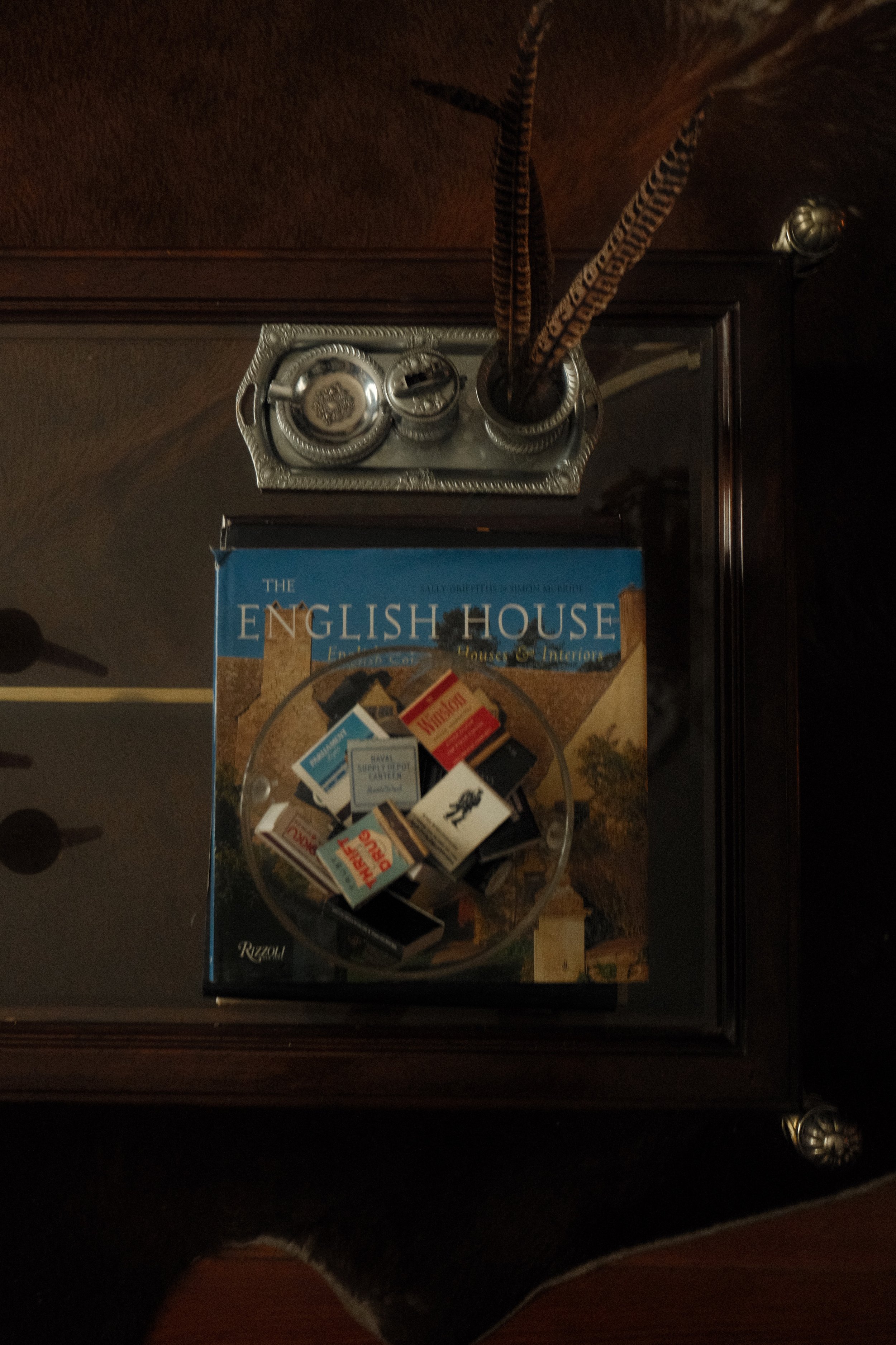 Top view of a silver-framed tray with incense sticks and ashtray, placed on a reflective glass surface. A magazine titled 'The English House' with miniature boxes on the cover is positioned below the tray.