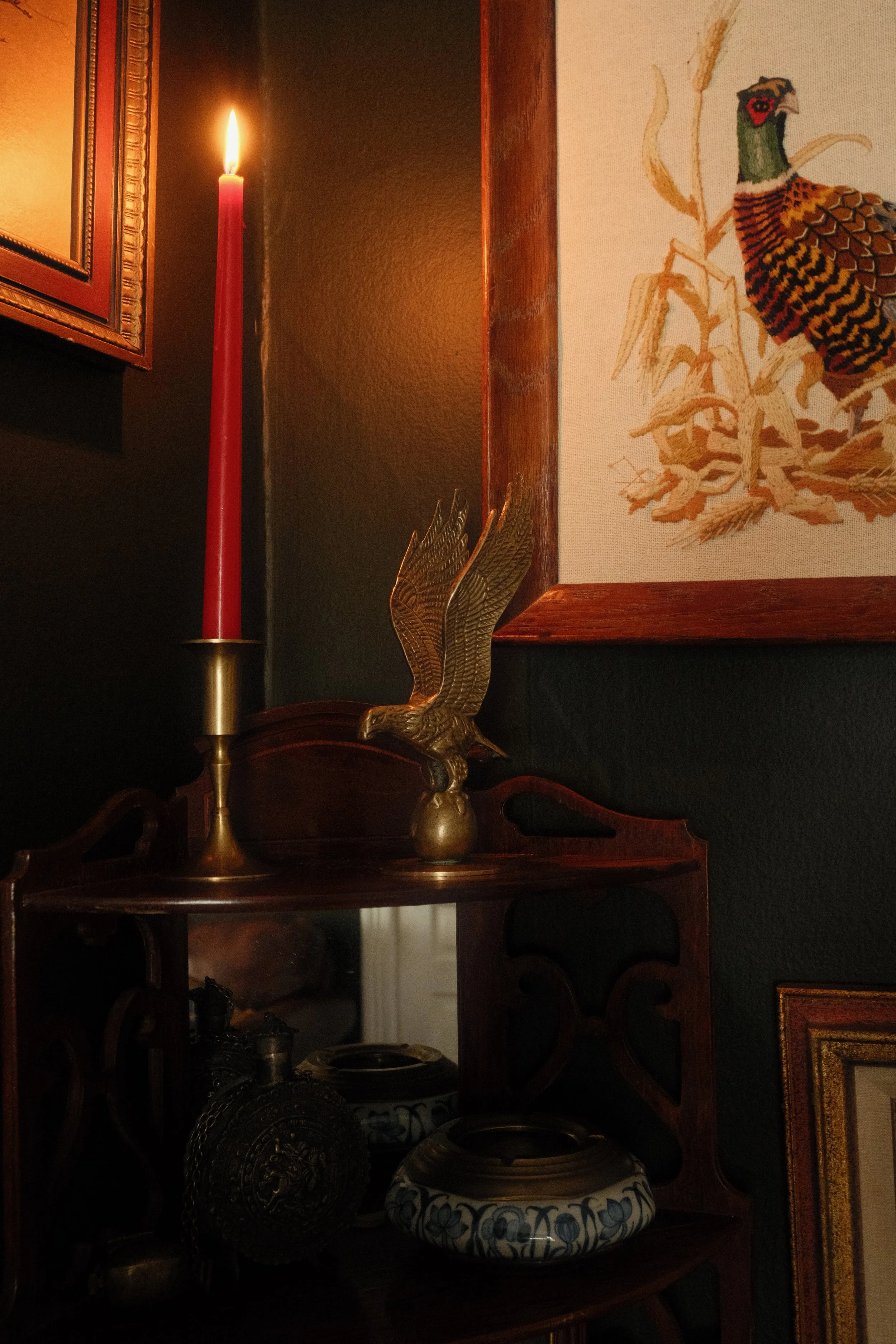 A lit red candle in a brass holder, decorative Asian-style brass eagle sculpture, sleeveless embroidered textile with a creature resembling a bird or dragon, and blue and white ceramic bowl with floral pattern, against a dark wall with framed artwork.