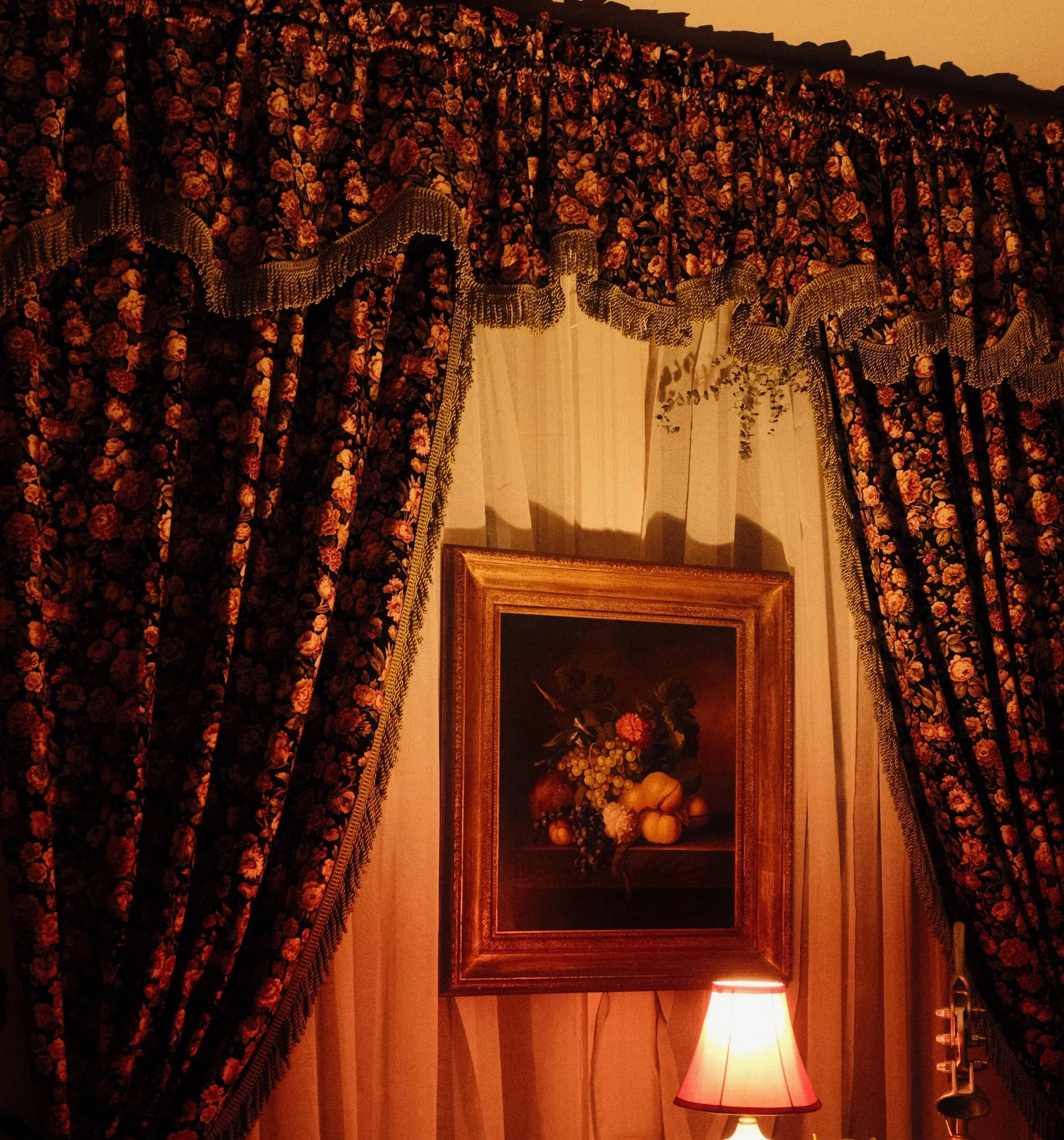 A wall with floral patterned curtains in dark colors and a sheer white curtain underneath, a framed still life painting of fruit and flowers, a table lamp with a pinkish shade, and a small decorative lamp next to a silver-colored object.