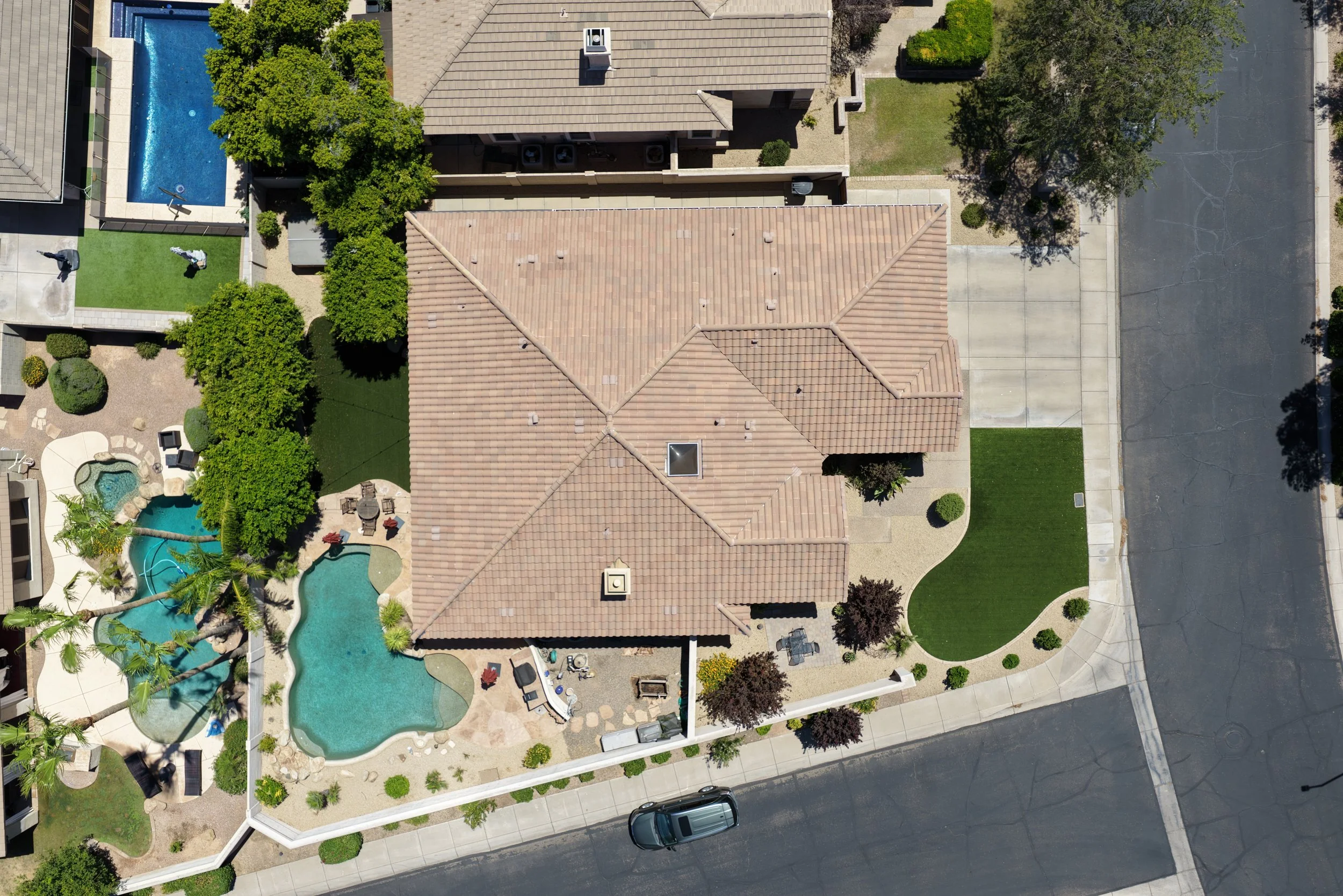 An aerial view of a residential backyard showing a swimming pool, a patio with outdoor furniture, trees, green lawns, and neighboring houses with tiled roofs and driveways.