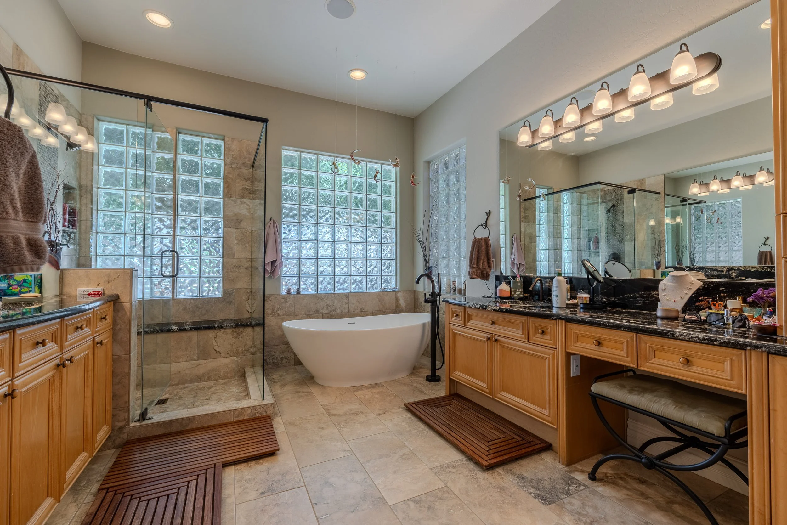 A large bathroom with a bathtub, glass-enclosed shower, wooden cabinetry, and large glass block windows. The room has beige tiled floors and walls, with two wooden mats and various personal items on the countertop.