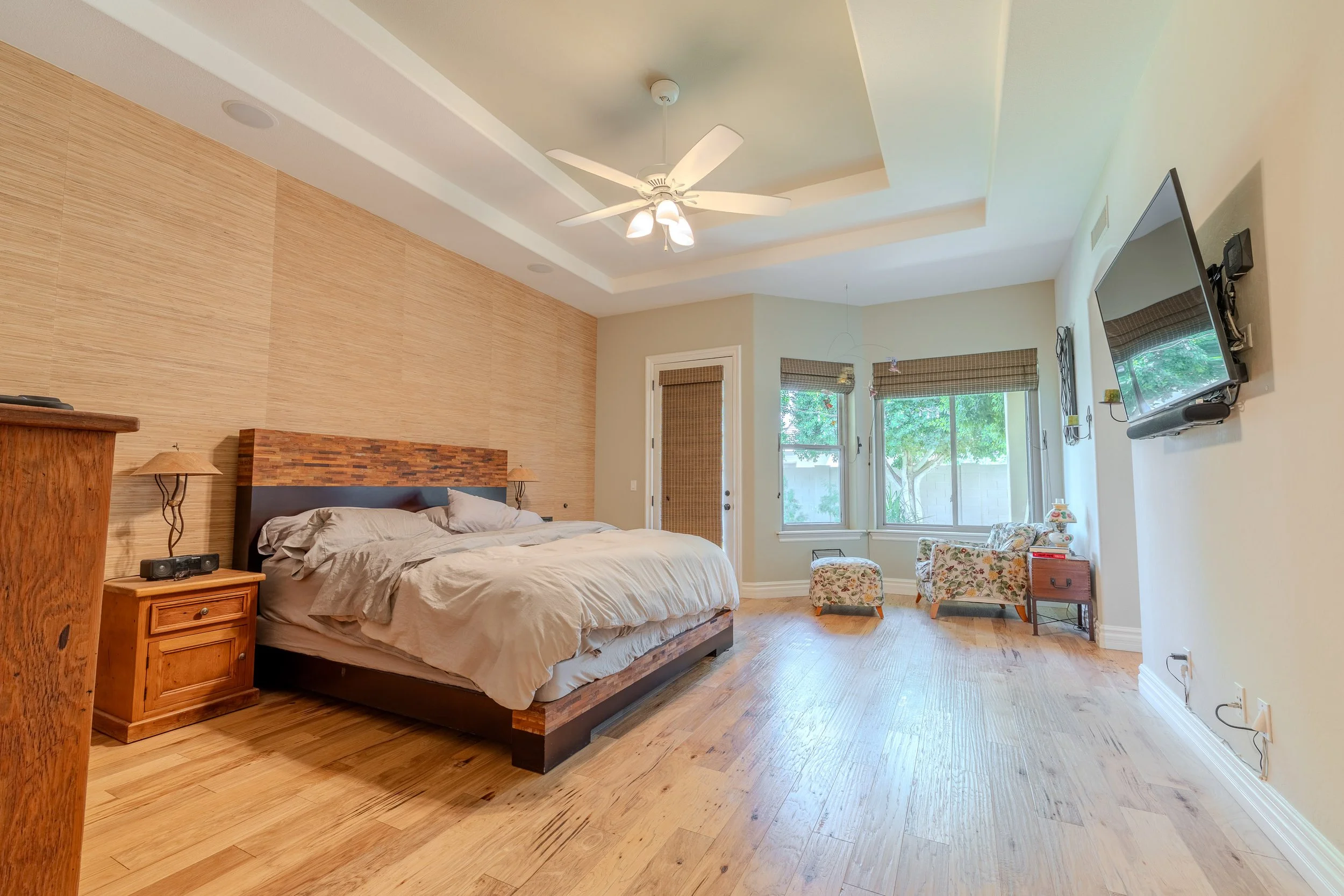 A bedroom with a wooden bed frame, beige bedding, wooden nightstand with a lamp, a wooden dresser, a wall-mounted flat screen TV, windows with blinds, a ceiling fan, and two floral armchairs.