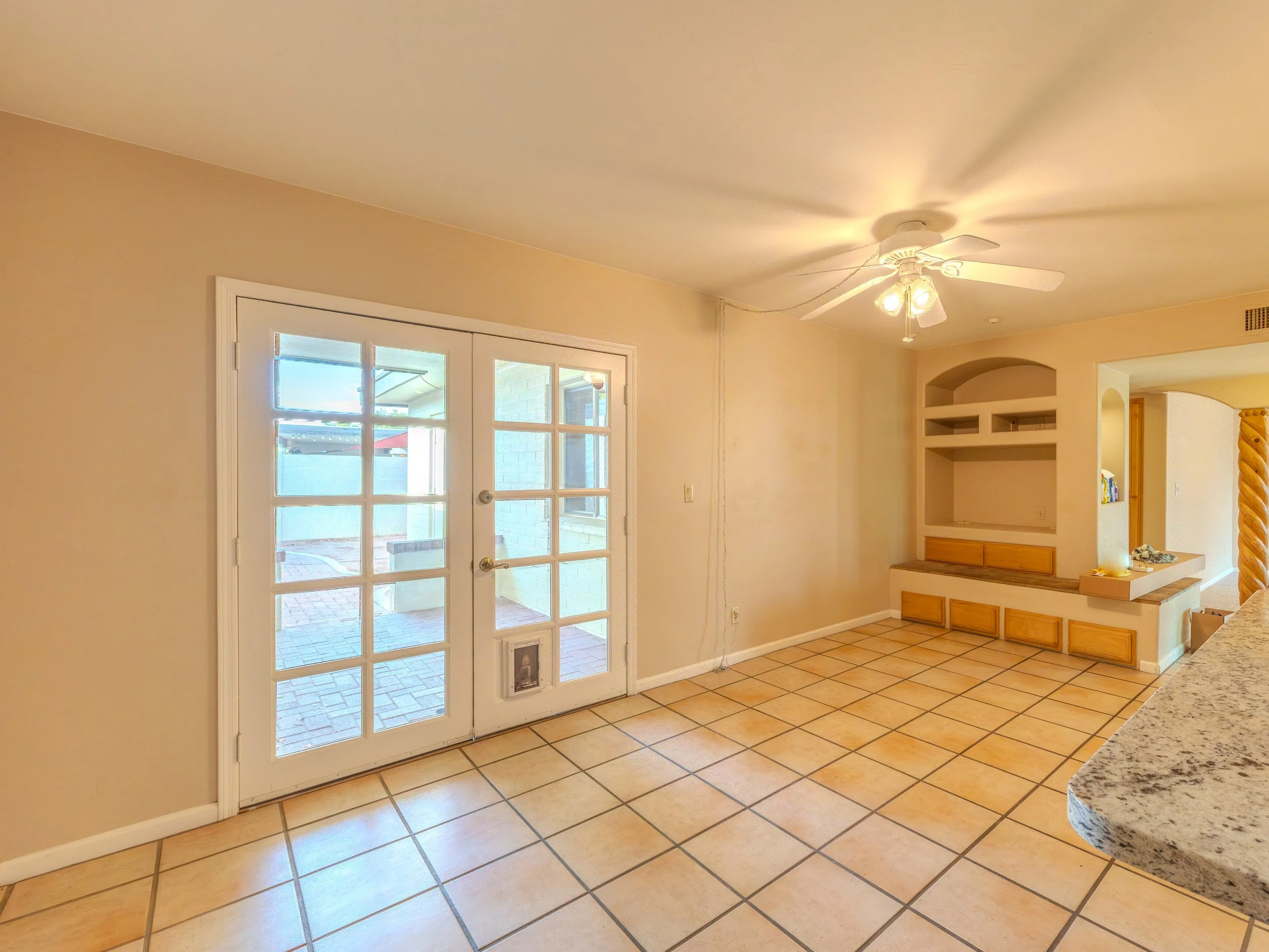 Interior view of a room with beige walls, ceramic tile floor, a ceiling fan with lights, and a glass-paneled door leading outside. There is a built-in shelf and a kitchen counter with wooden drawers and granite countertop visible.