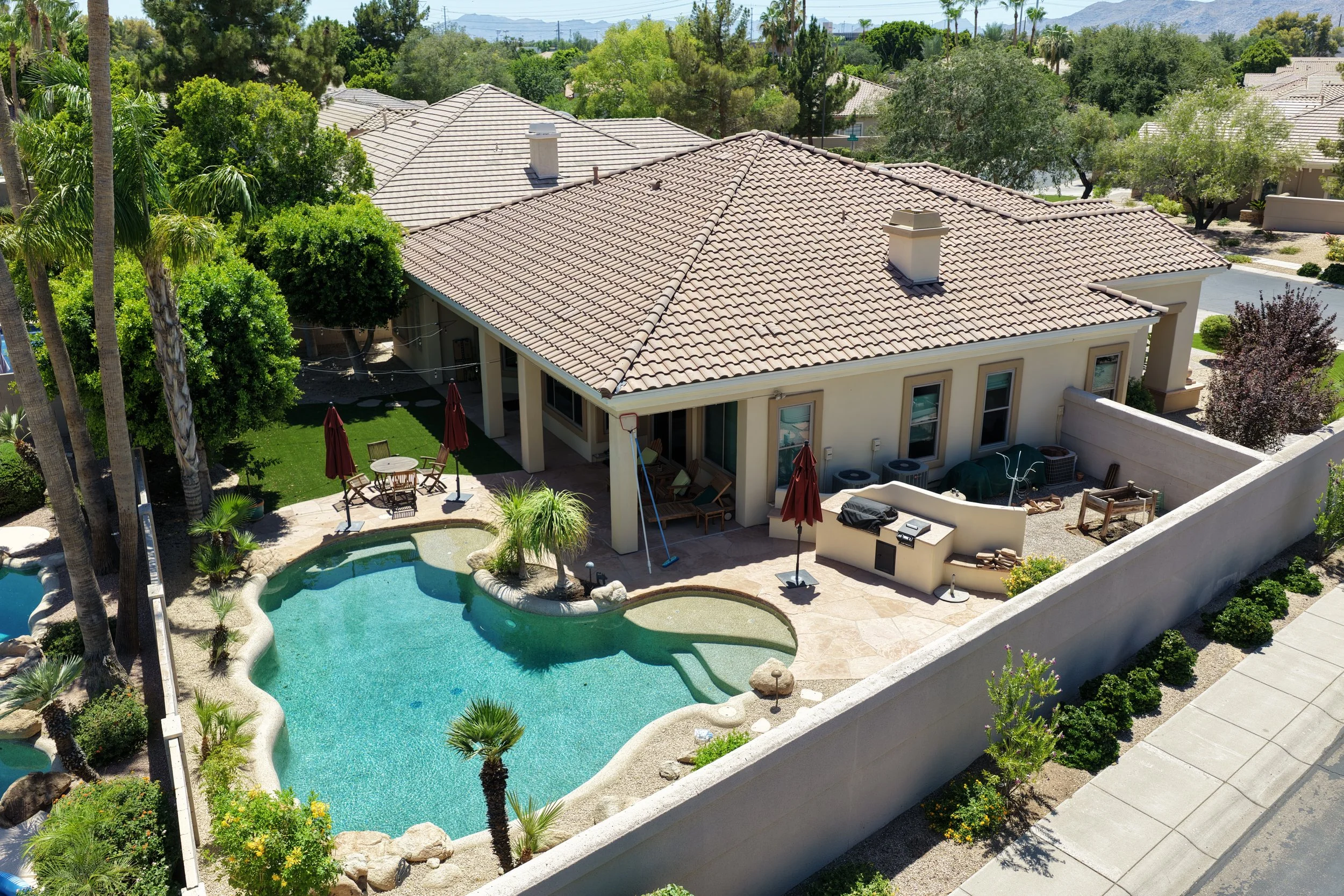 An aerial view of a backyard with a swimming pool and patio area, surrounded by trees and a house with a tiled roof, in a suburban neighborhood.