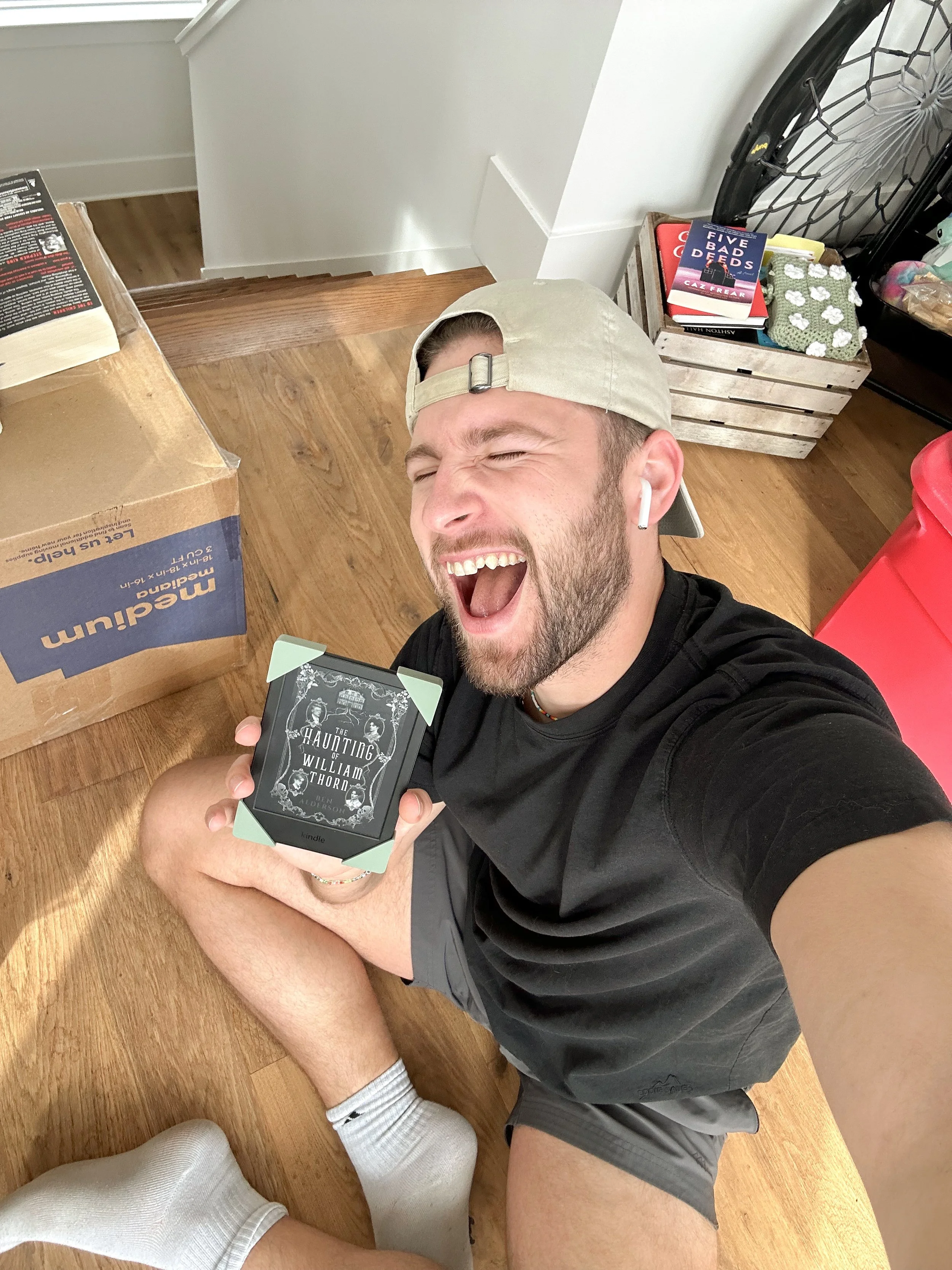 Young man with a beard and cap, sitting on the floor and smiling as he holds a Kindle e-reader with the book title 'The Haunting of William Thorn' visible on the screen.