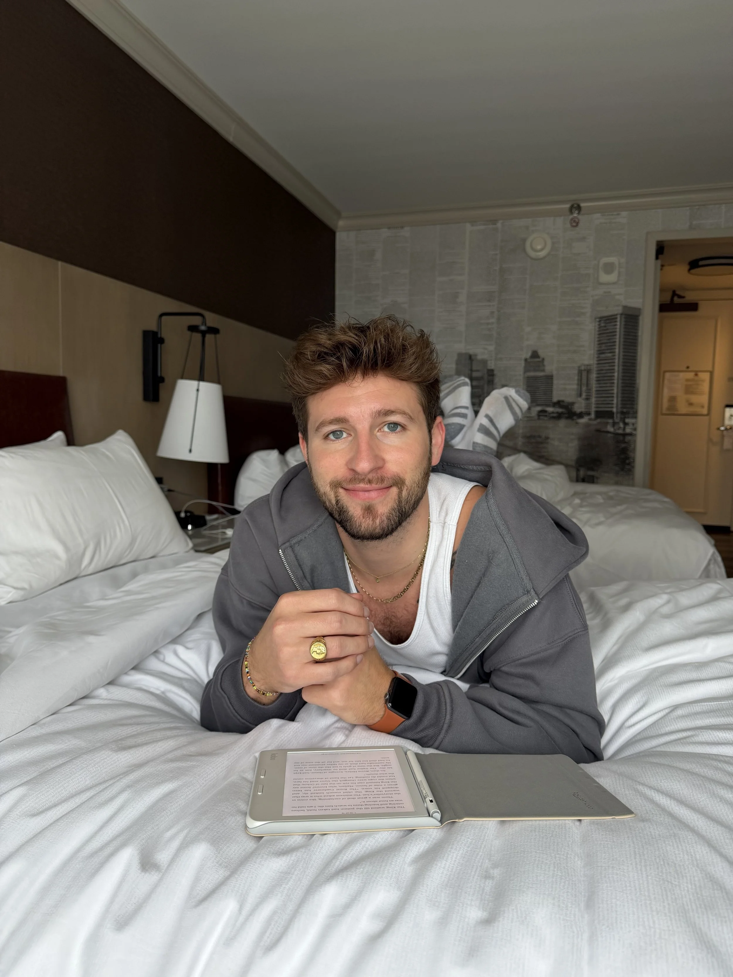 A young man with brown hair and blue eyes lying on a bed with white sheets, in a hotel room. He wearing a gray hoodie, a white t-shirt, and has jewelry including a gold ring, a bracelet, and a necklace. There is an  with text on the screen in front of him.