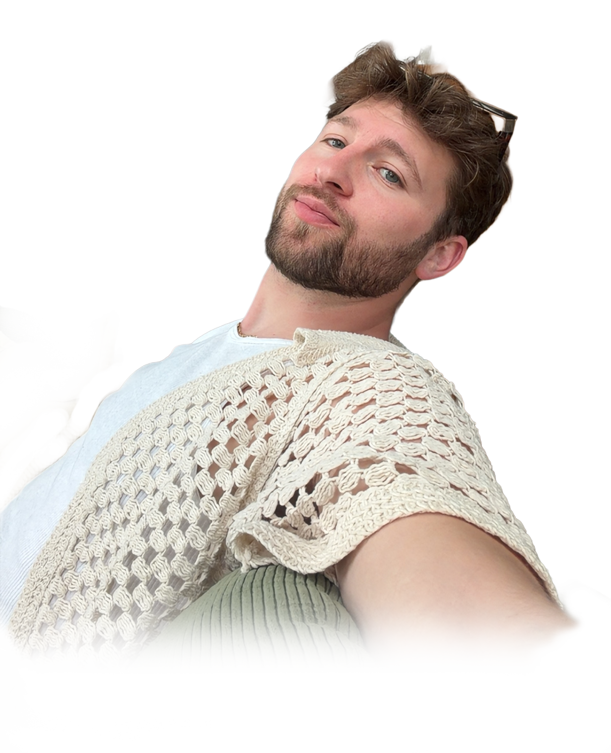A young man with curly brown hair, a beard, and blue eyes is smiling softly while reclining. He's wearing a white shirt and a cream-colored, crocheted cardigan, with sunglasses perched on his head.