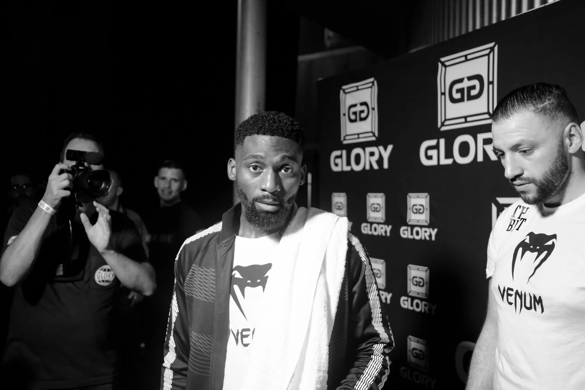 Two esports players with towels around necks at a post-match interview, with a backdrop that reads "GLORY" and features logos, one player with dark curly hair and beard, the other with light hair and beard, and a cameraman filming them.