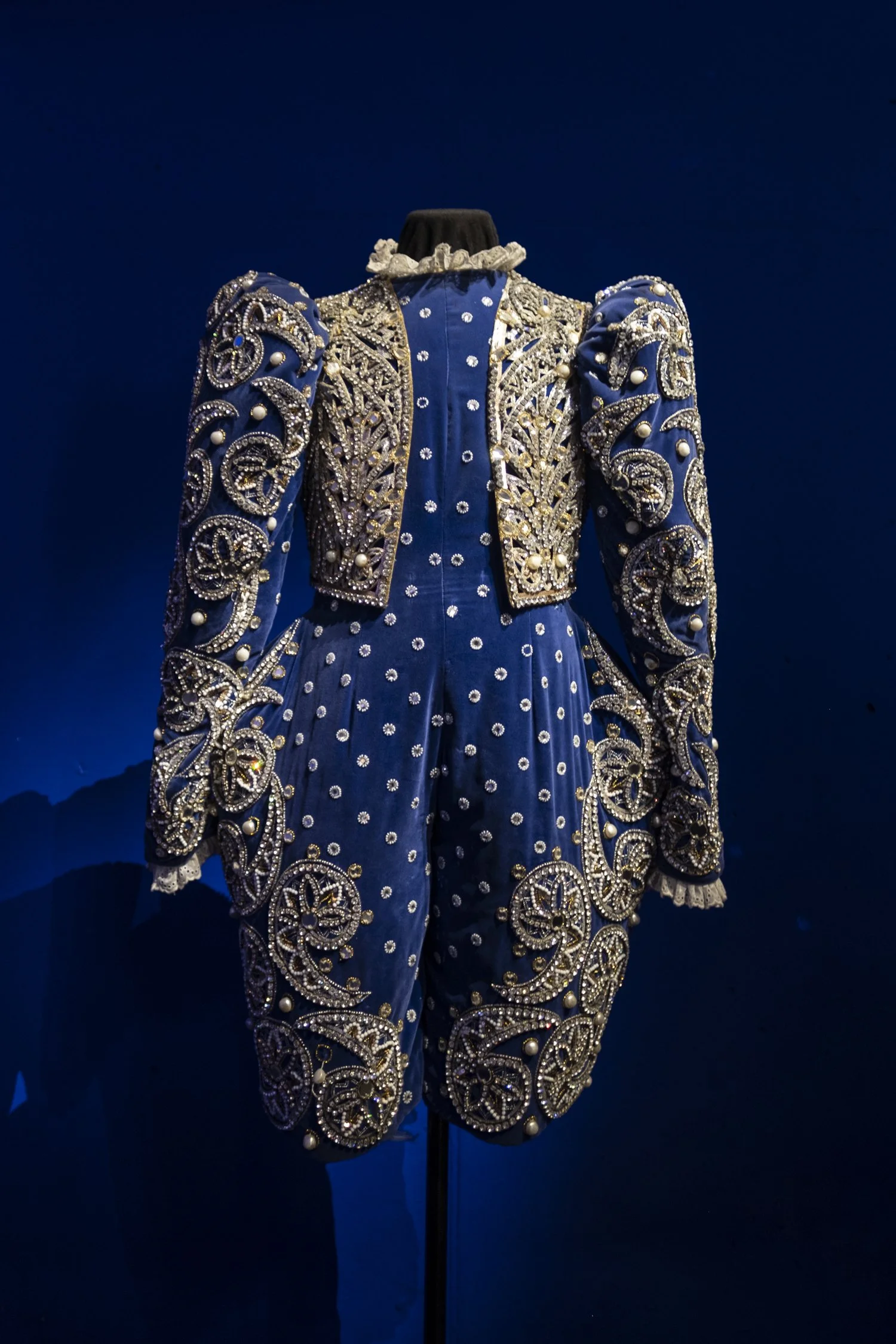 A blue ornate costume with gold and pearl embellishments, displayed on a mannequin against a dark blue background. Swarovski.