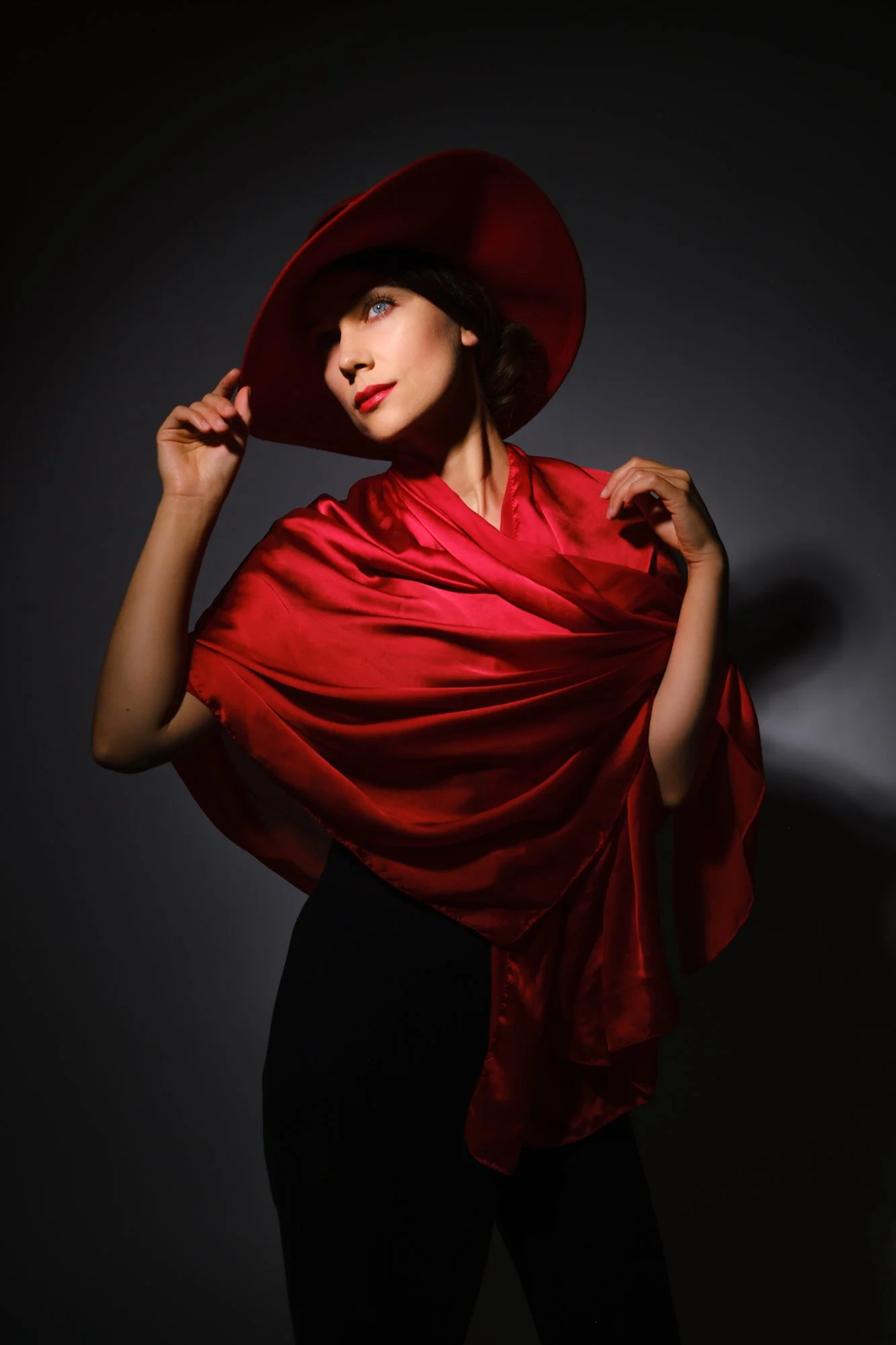 A woman with blonde hair and blue eyes. She is dressed in a red silk shawl and wide-brimmed red hat, posing against a dark background.
