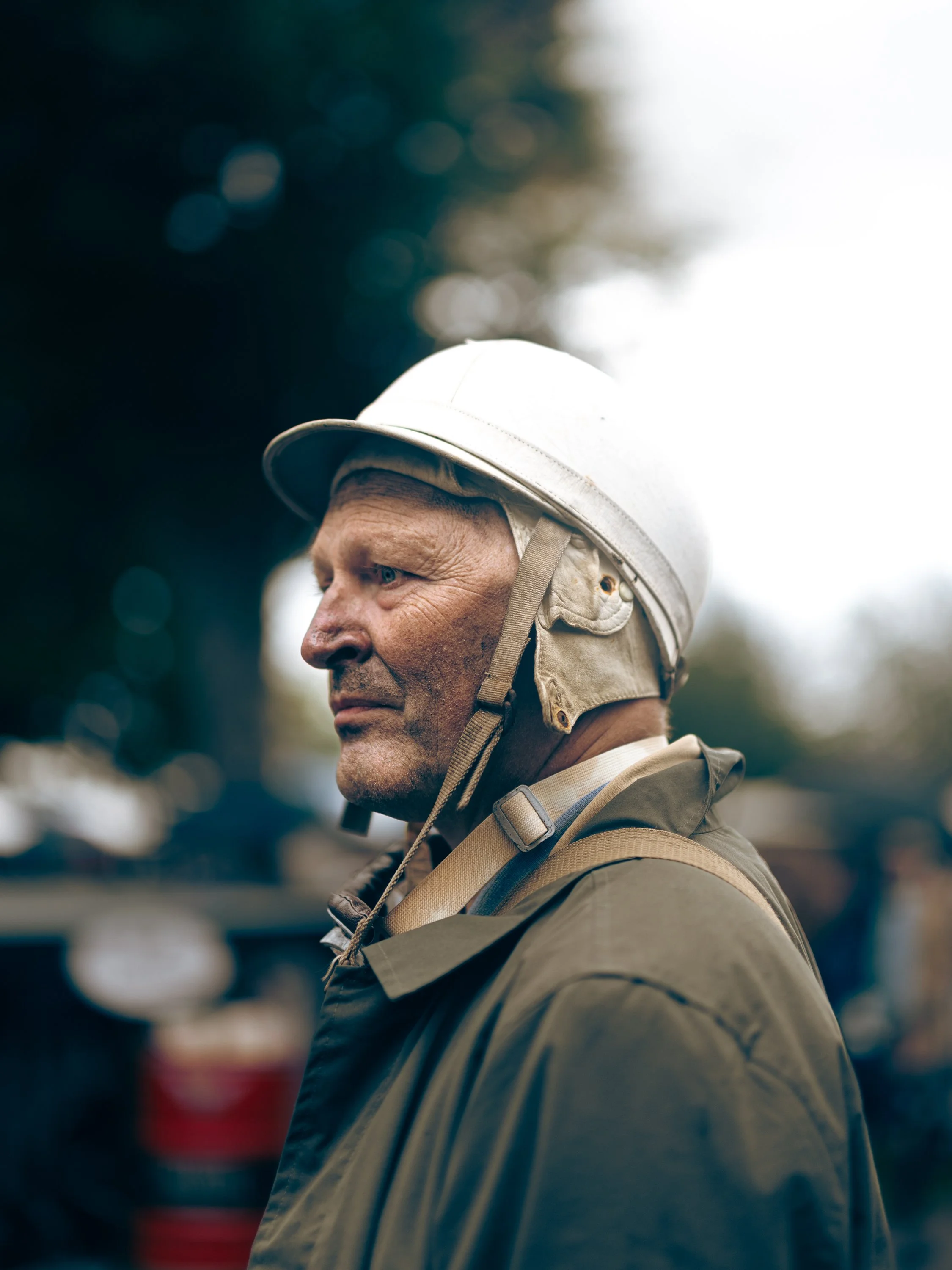 Close-up of a man wearing a beige helmet and outdoor clothing, looking off to the side, with blurred figures and trees in the background.