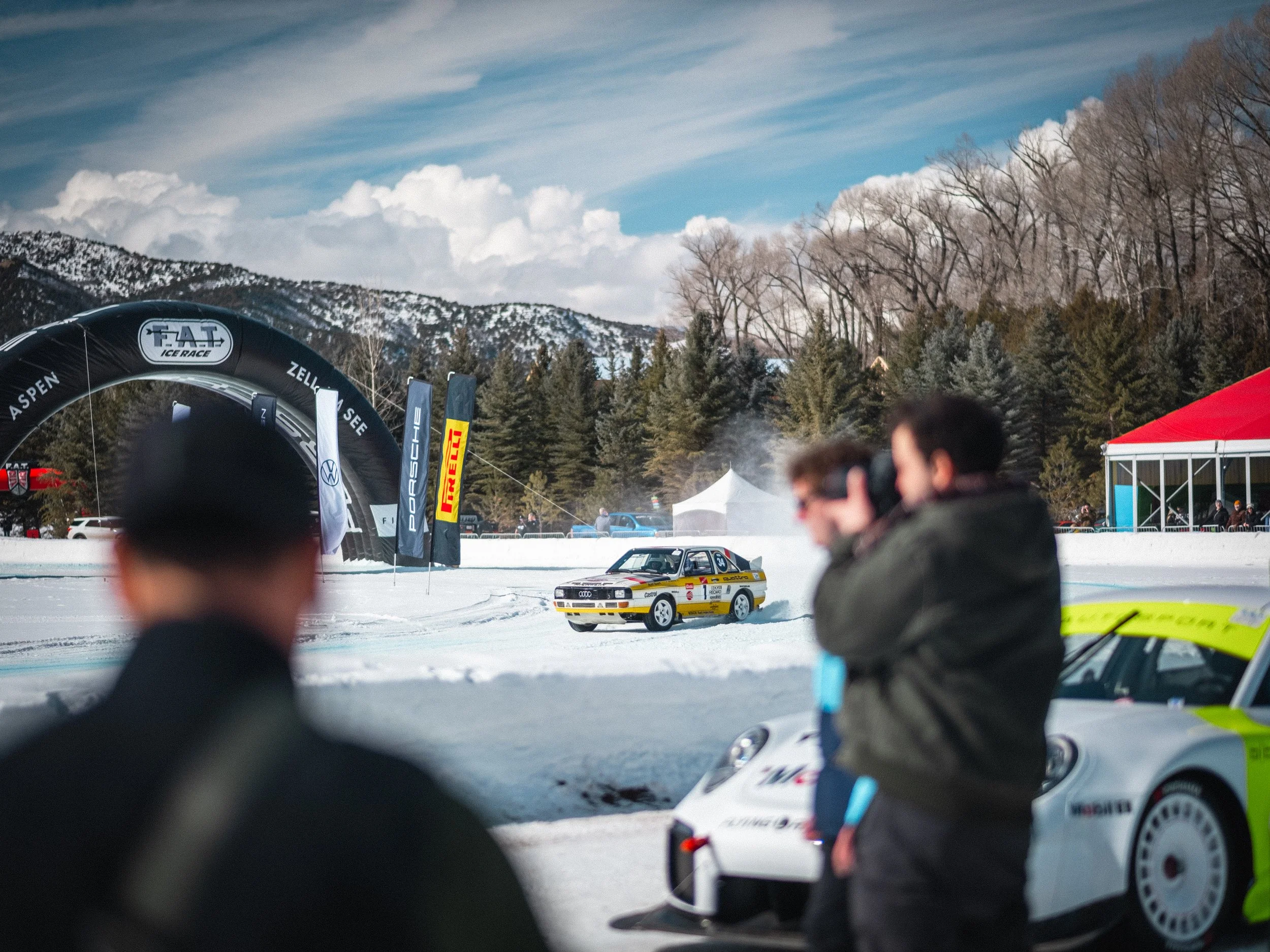Race car on snow at an outdoor racing event with mountains, trees, and event banners in the background. Audi Quattro rally car, Group B rally, Fat Ice Race