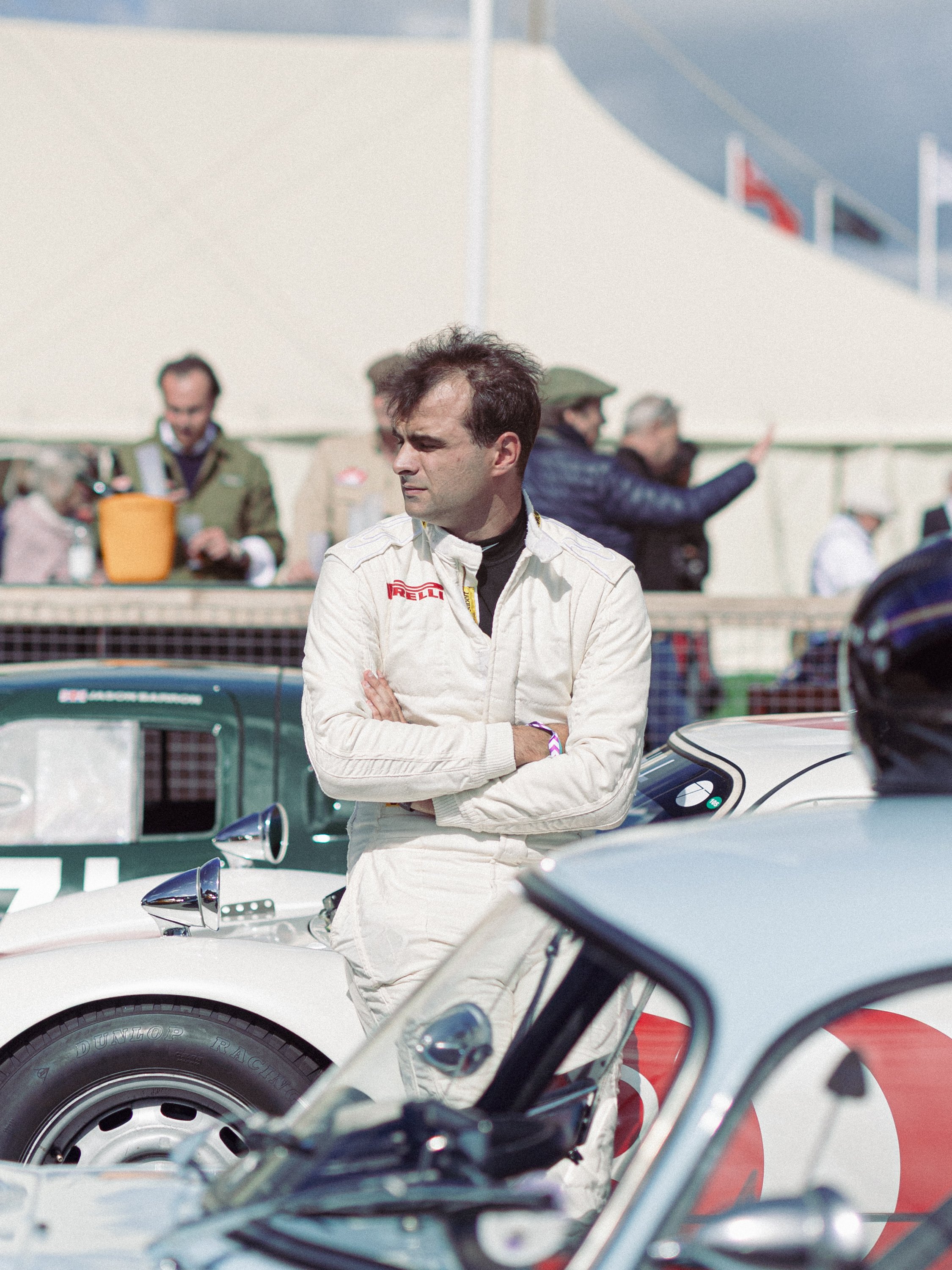 A man in a white racing suit with a Pirelli logo stands with arms crossed, surrounded by racing cars and other people at a racing event. Goodwood Revival.
