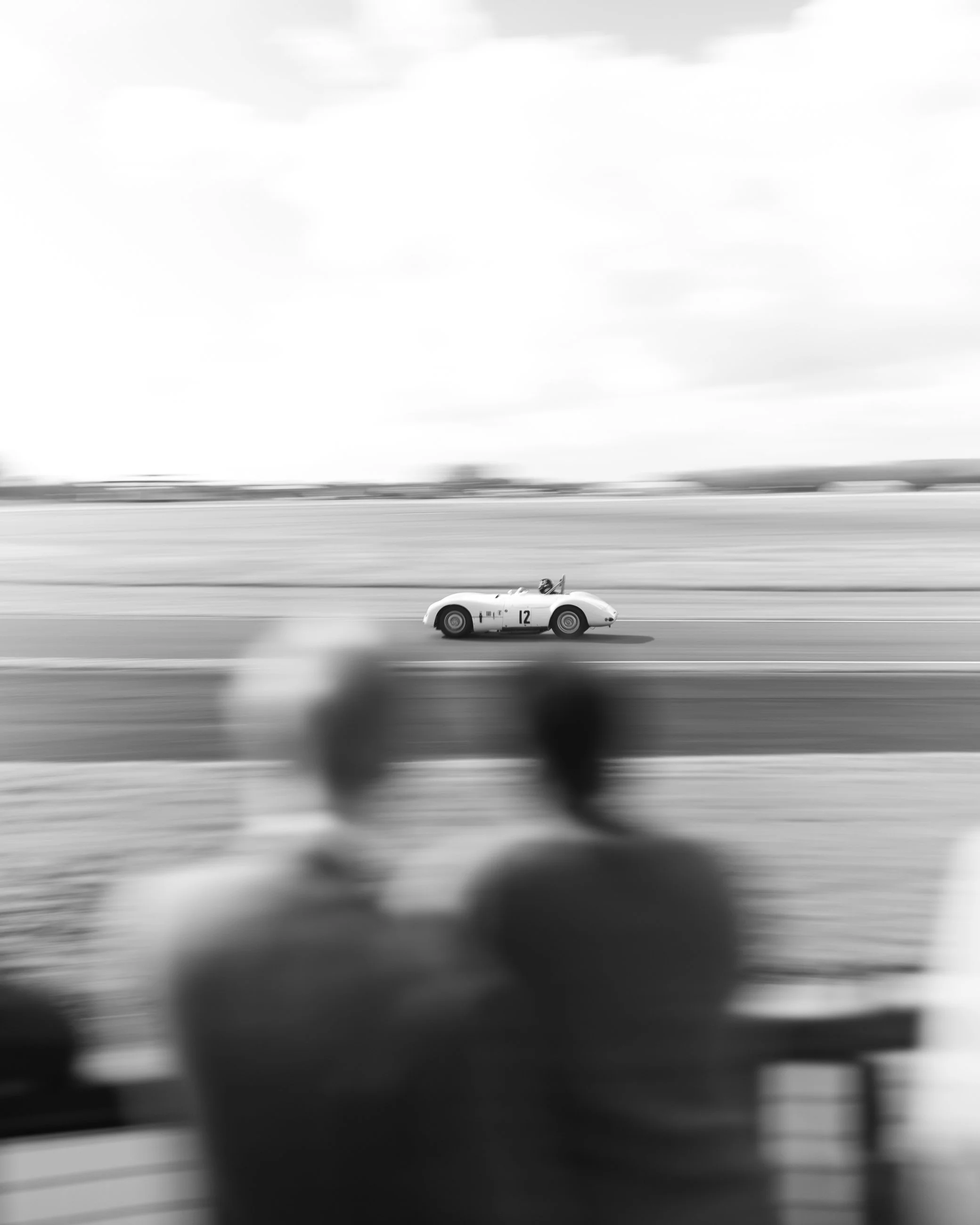 Black and white photo of a vintage race car with the number 12, speeding along a track blurred to show motion, with a driver in the seat and a distant background of sky and landscape. Goodwood Revival.