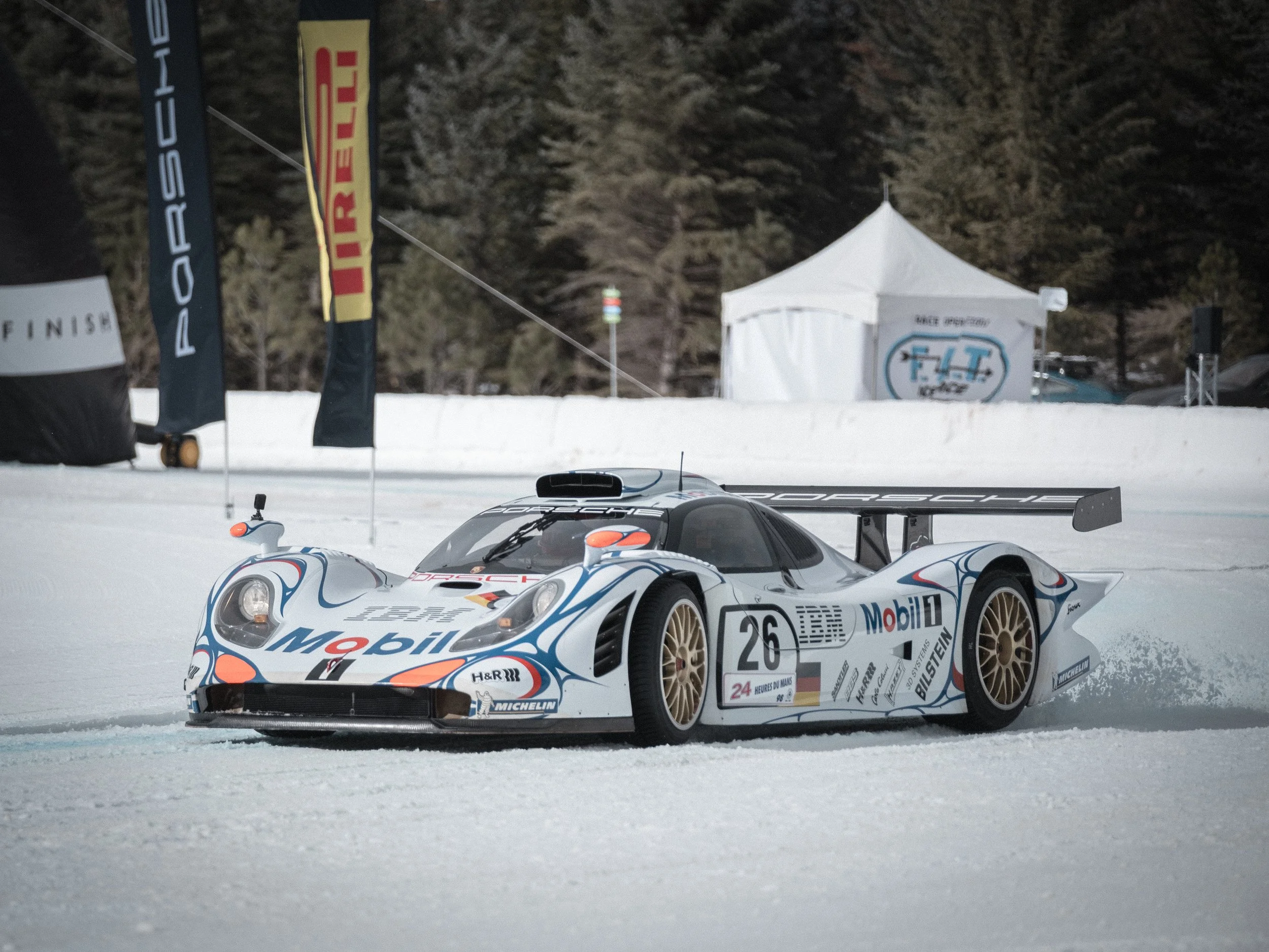 A racing car on snow-covered ground at night with a white tent and trees in the background. A race car moving on a snowy track with trees and buildings in the background. Porsche LeMans race car Fat Ice Race
