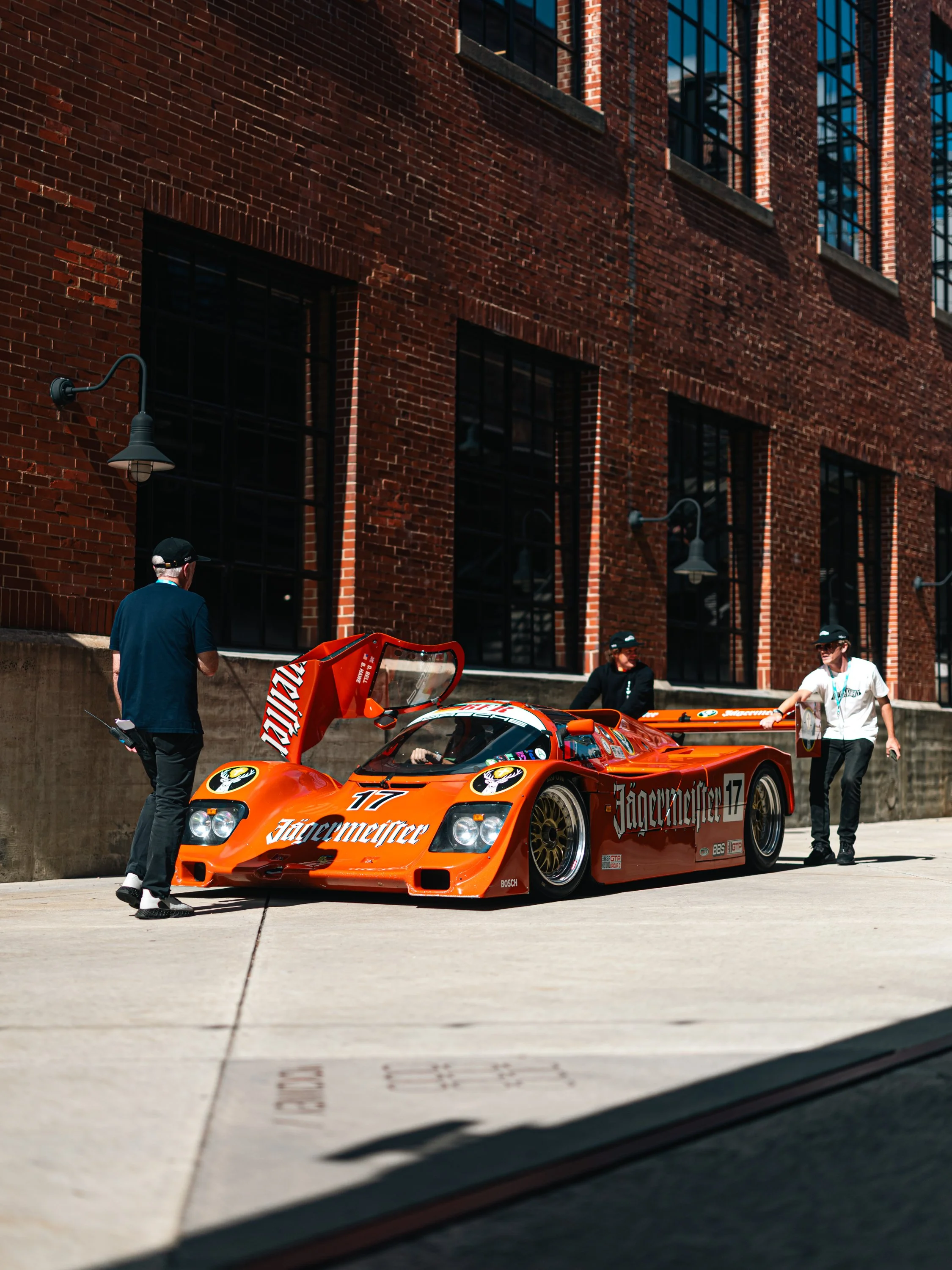 Orange race car with 'Jägermeister' branding and the number 17 on it, parked outside a brick building, with three men nearby, one in a black cap and blue shirt, and two others in black and white helmets.