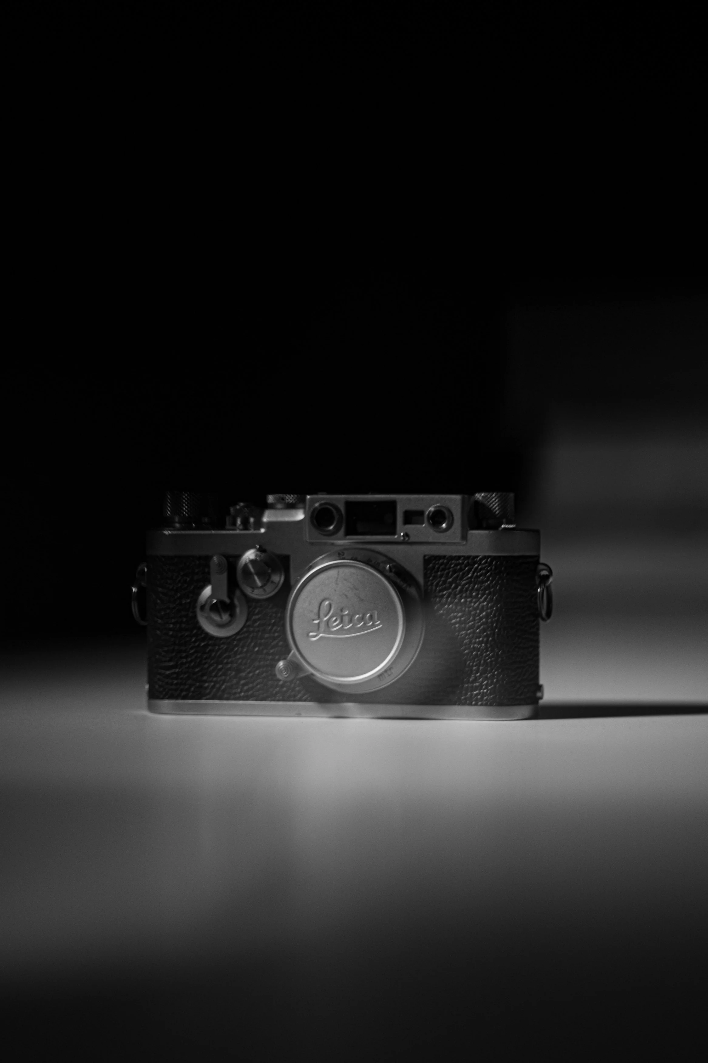 Vintage Leica camera with a textured black body and silver top, placed on a flat surface, centered in a dark setting.