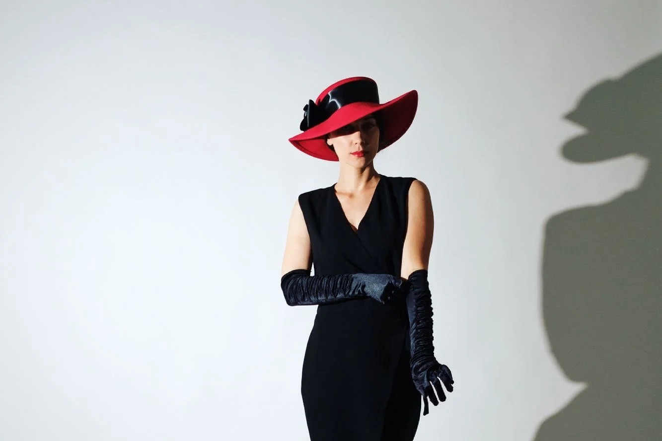A woman in a black dress and long black gloves wearing a large red and black wide-brimmed hat casting a shadow on a plain white background.