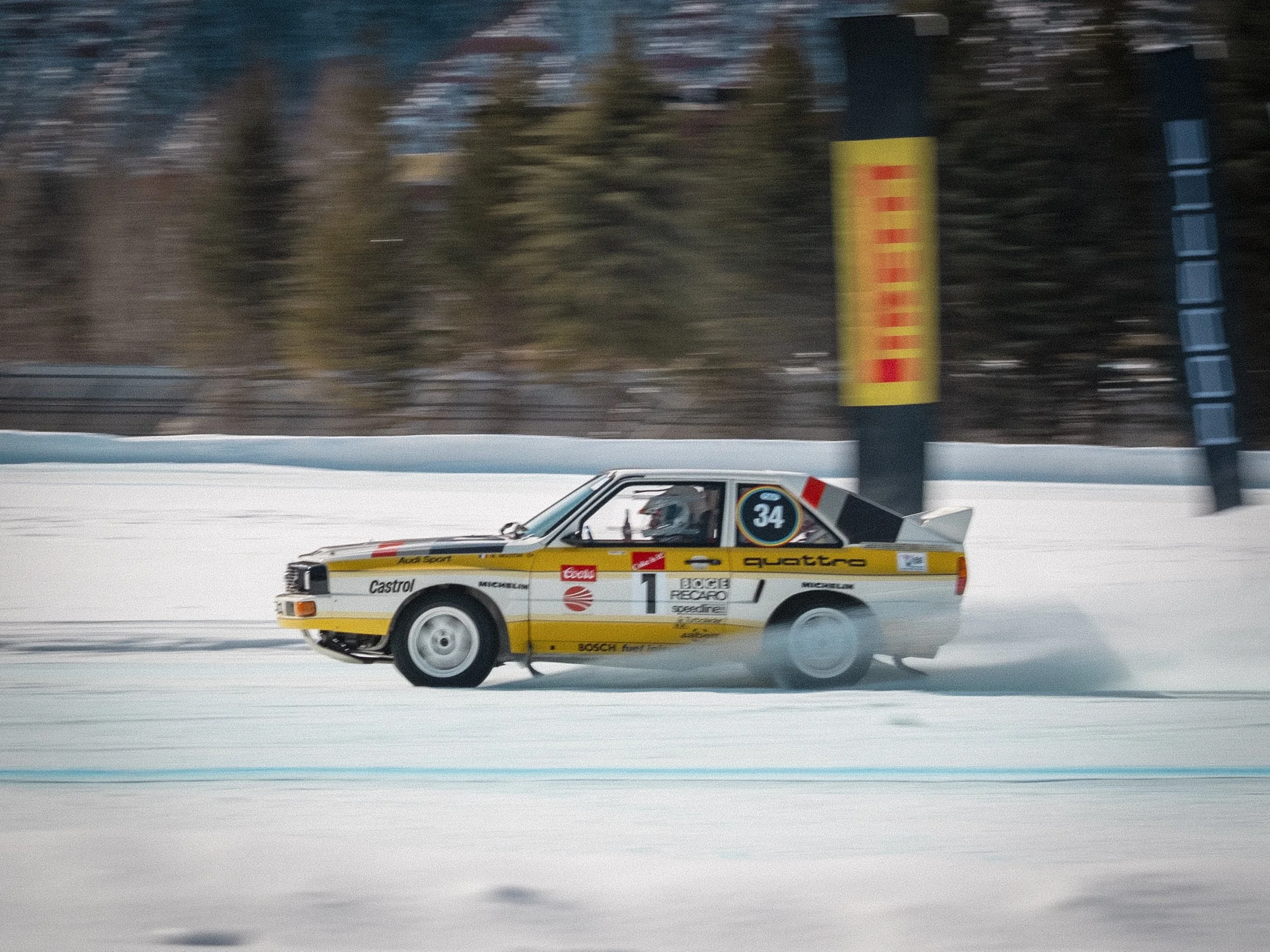 A vintage rally car racing on a snowy track, with a background of trees and a yellow banner. Audi Quattro rally car, Group B rally, Fat Ice Race, Aspen Colorado