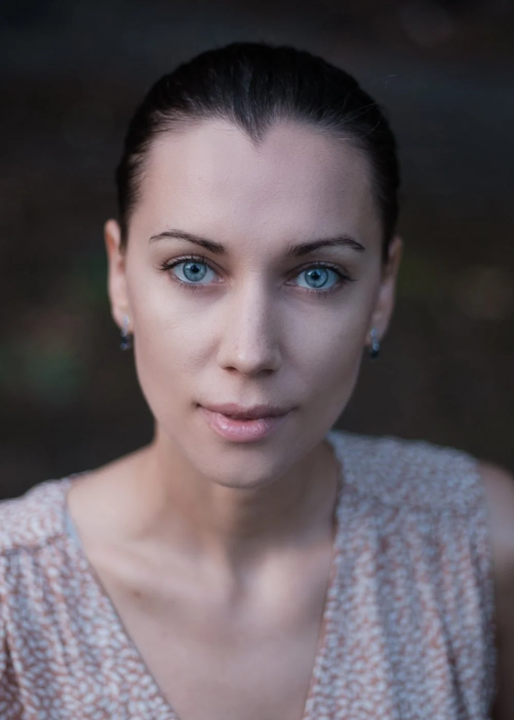 Close-up of a woman with blue eyes, dark hair tied back, wearing earrings and a patterned top, outdoors in natural light.