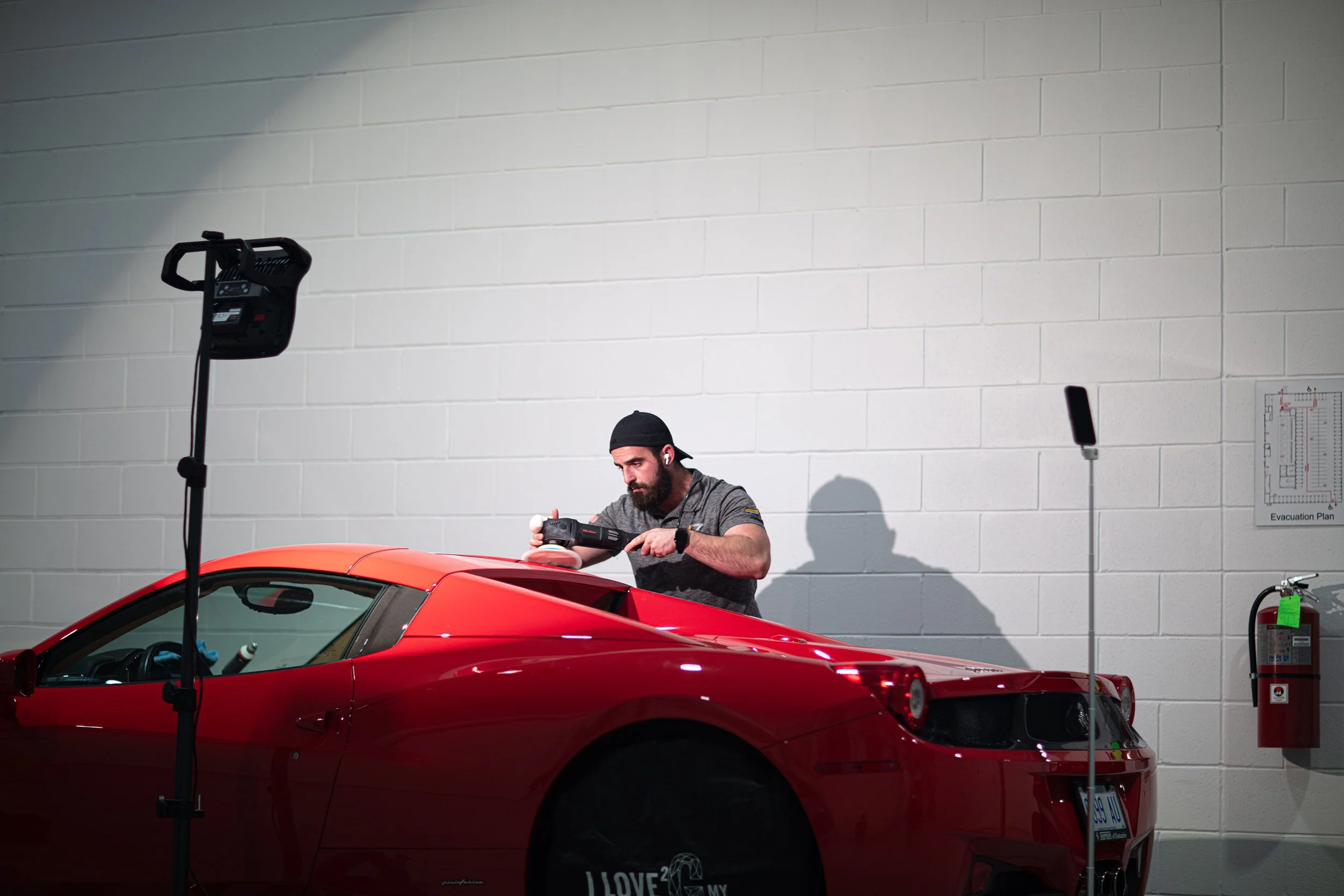 A man wearing a black cap and earbuds is working on a red sports car inside a garage. There is a large light stand and an evacuation plan on the wall behind him.