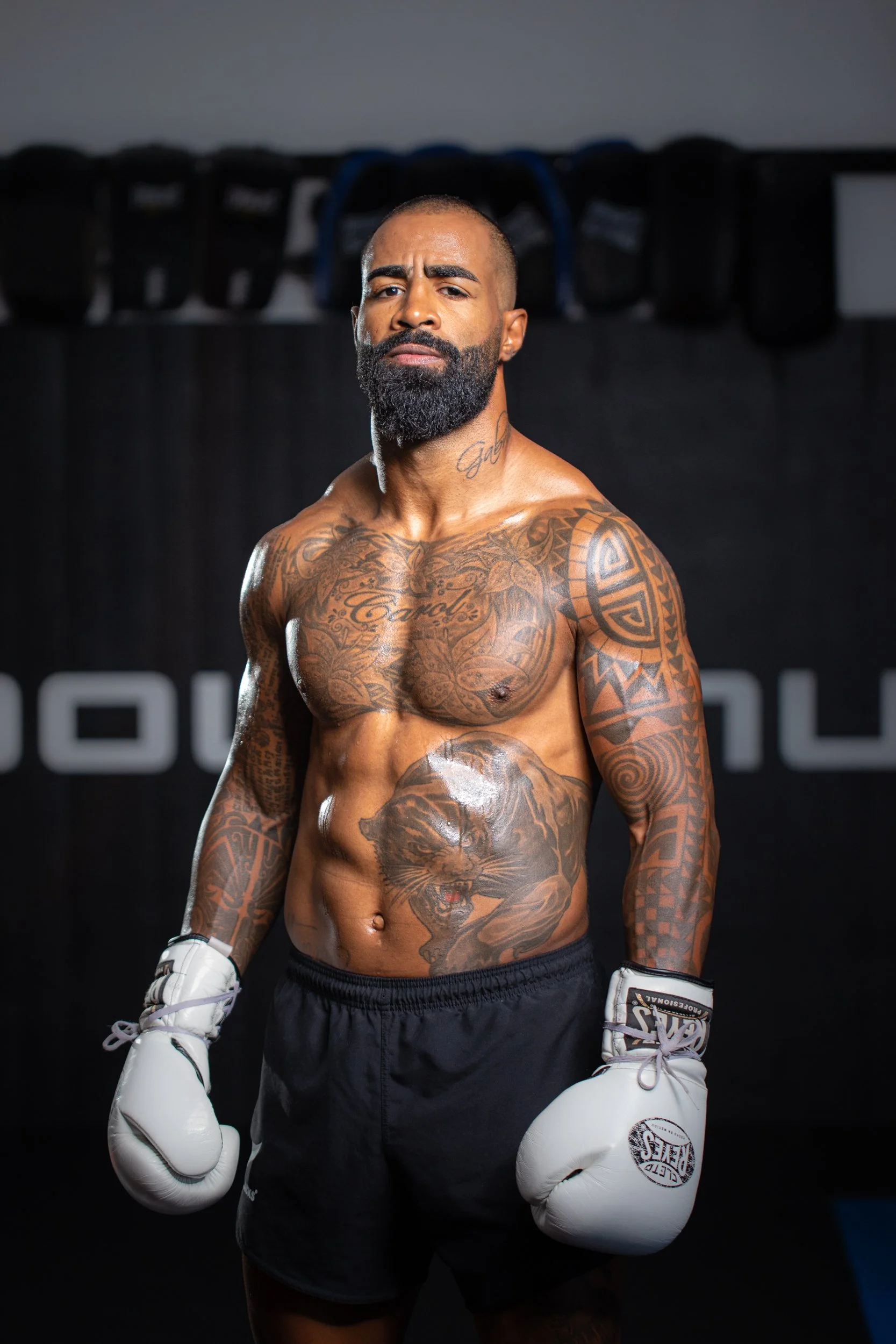 A muscular man with tattoos, wearing boxing gloves and shorts, standing in a gym with a black background. Cosmo Alexandre, muay thai