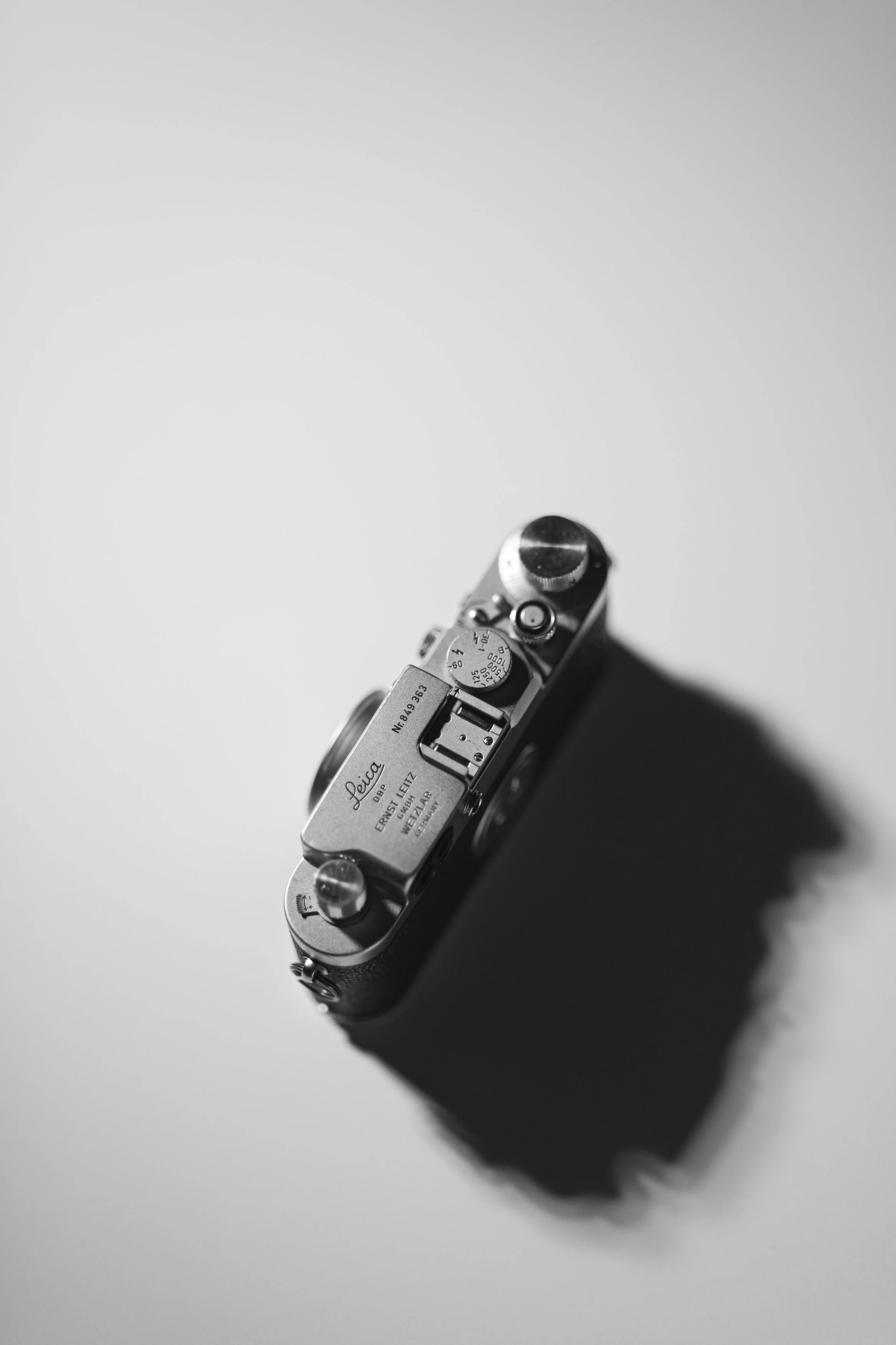A black and white photograph of a vintage Leica camera viewed from above against a plain, light-colored background.