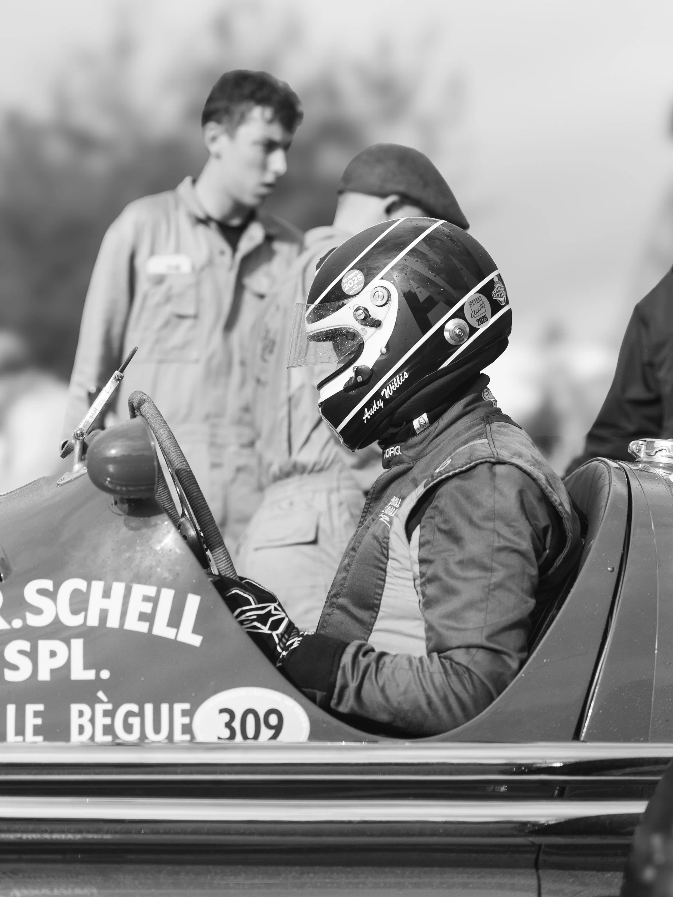 Close-up of a race car driver in a helmet, seated in a racing car with partial view of the car's windshield and body, and a person standing nearby. The car has French text and circular number decals. Goodwood Revival.