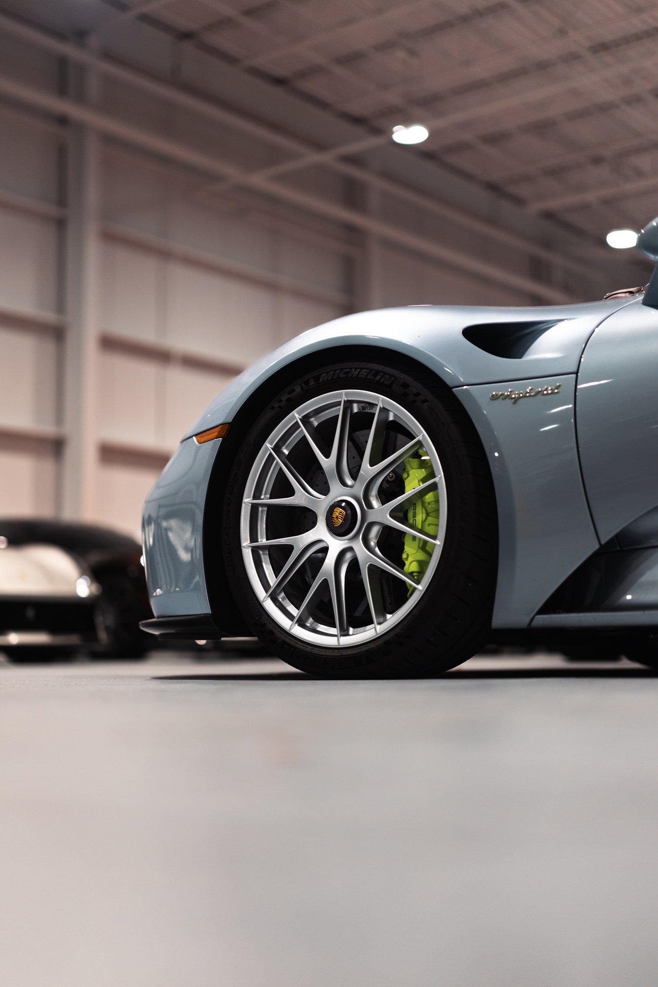 Close-up of a silver sports car with green brake calipers and a Porsche emblem, parked indoors with other cars in the background. Porsche 918
