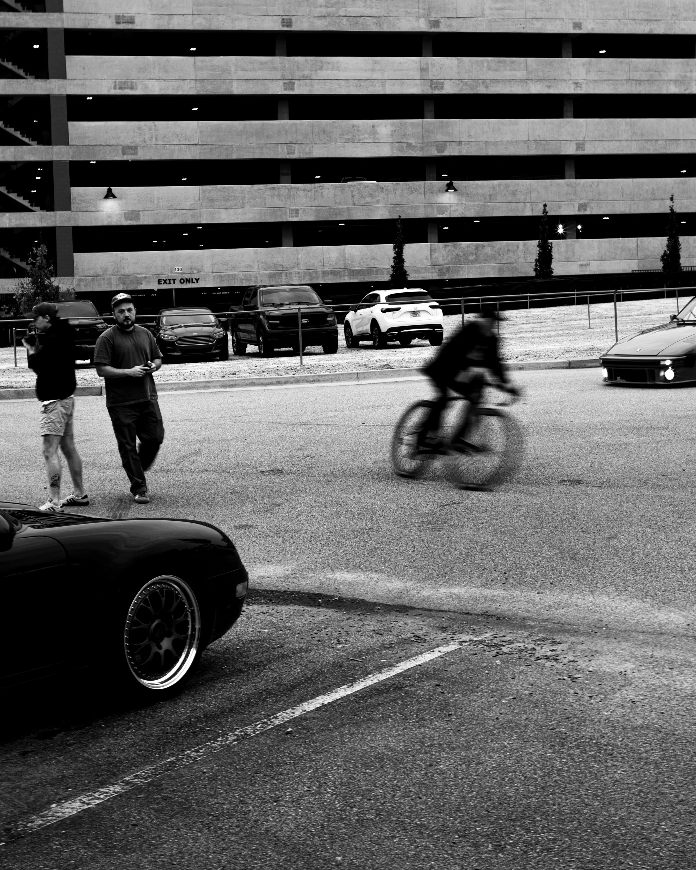 Black-and-white street scene with pedestrians, a blurred cyclist, parked cars, and a concrete parking garage marked “EXIT ONLY.”