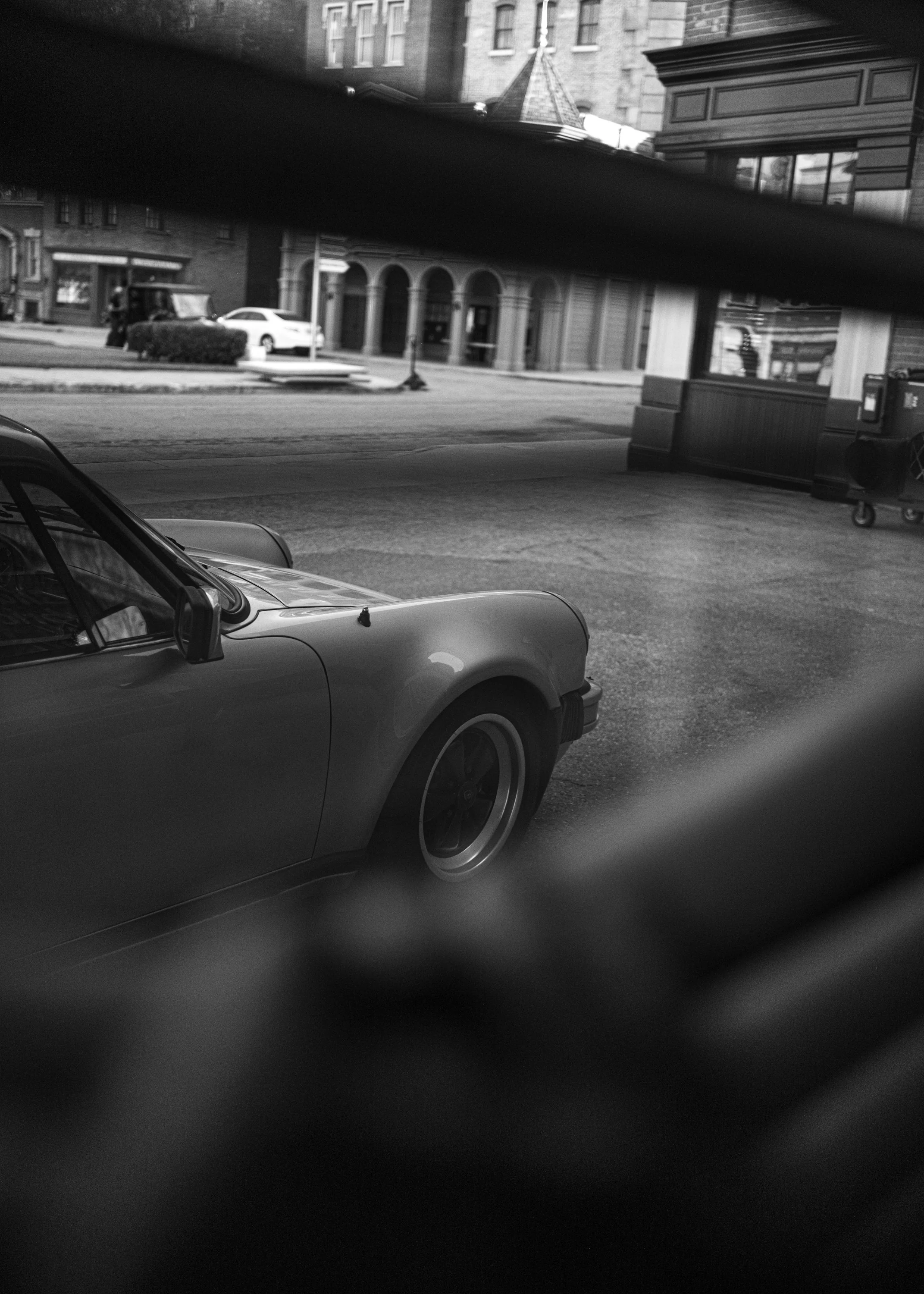 Black and white photo of a car parked on a street, with buildings and a sidewalk in the background, view partially obstructed by a dark horizontal object.