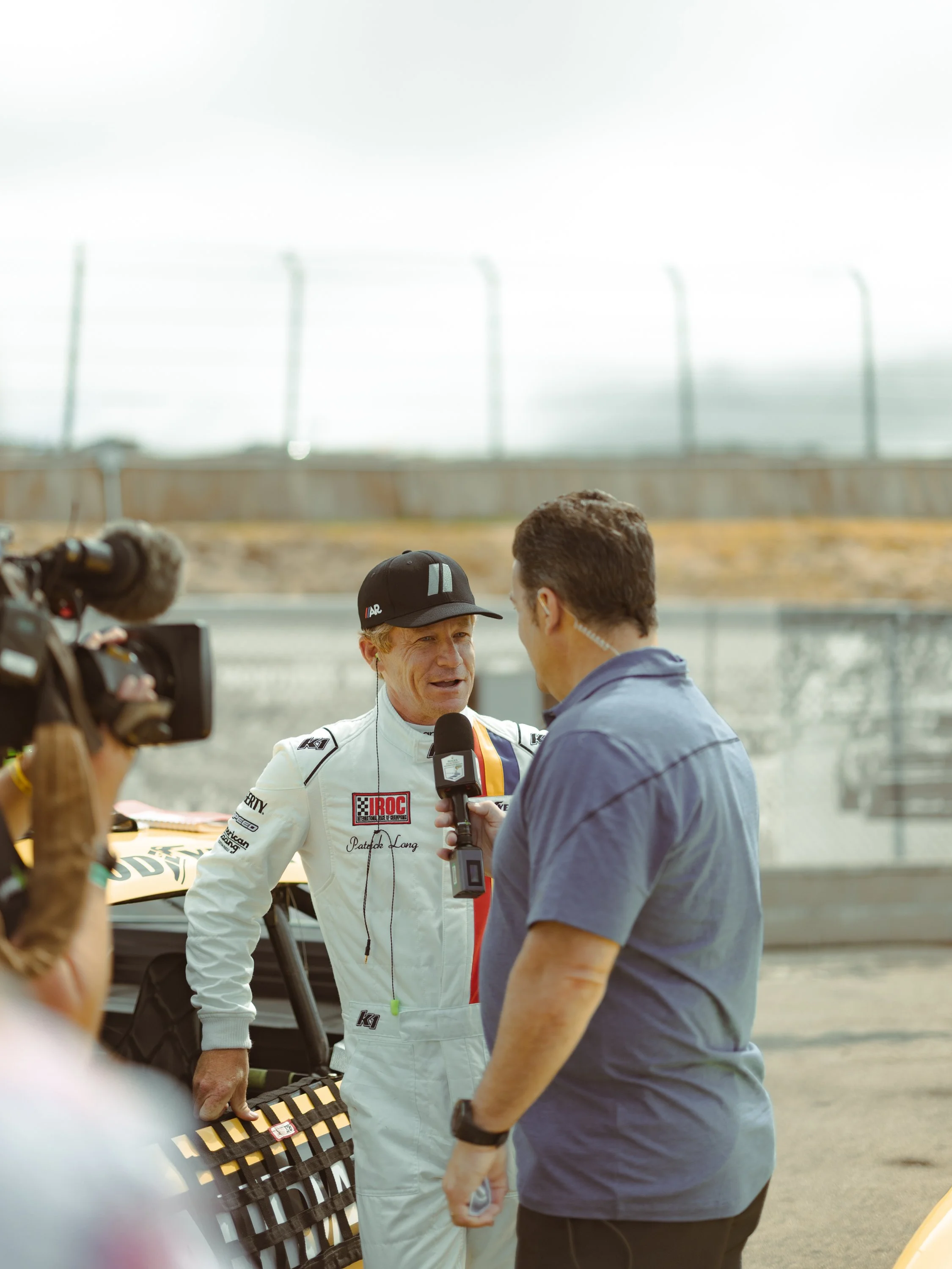Race car driver in white racing suit being interviewed by a microphone, with a man in a blue shirt and holding a notebook, standing next to a yellow race car on a racetrack. Patrick Long