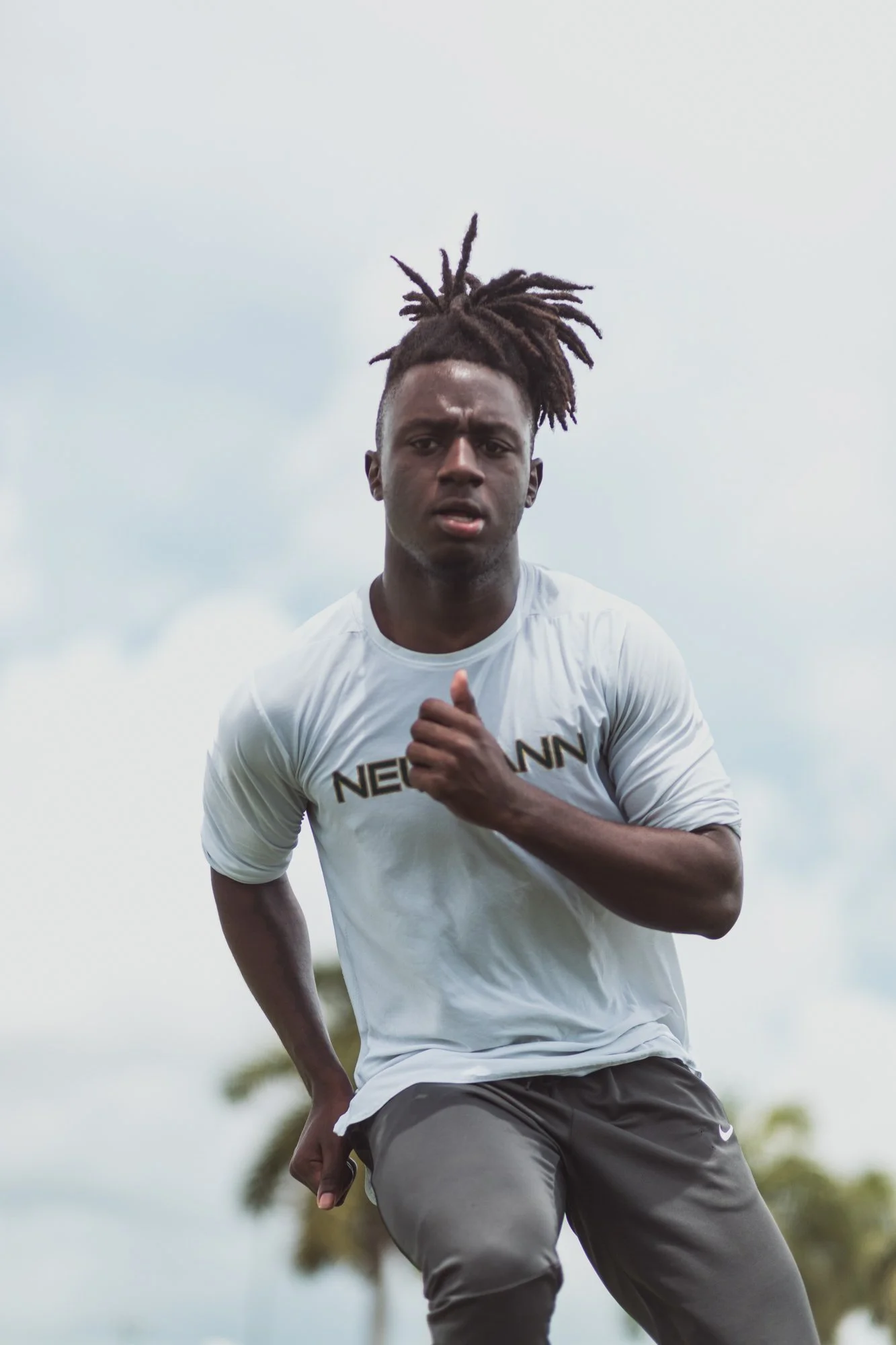 Young man jogging outdoors, wearing a white Nike T-shirt and gray Nike pants, with a serious expression and focused gaze, against a cloudy sky background.