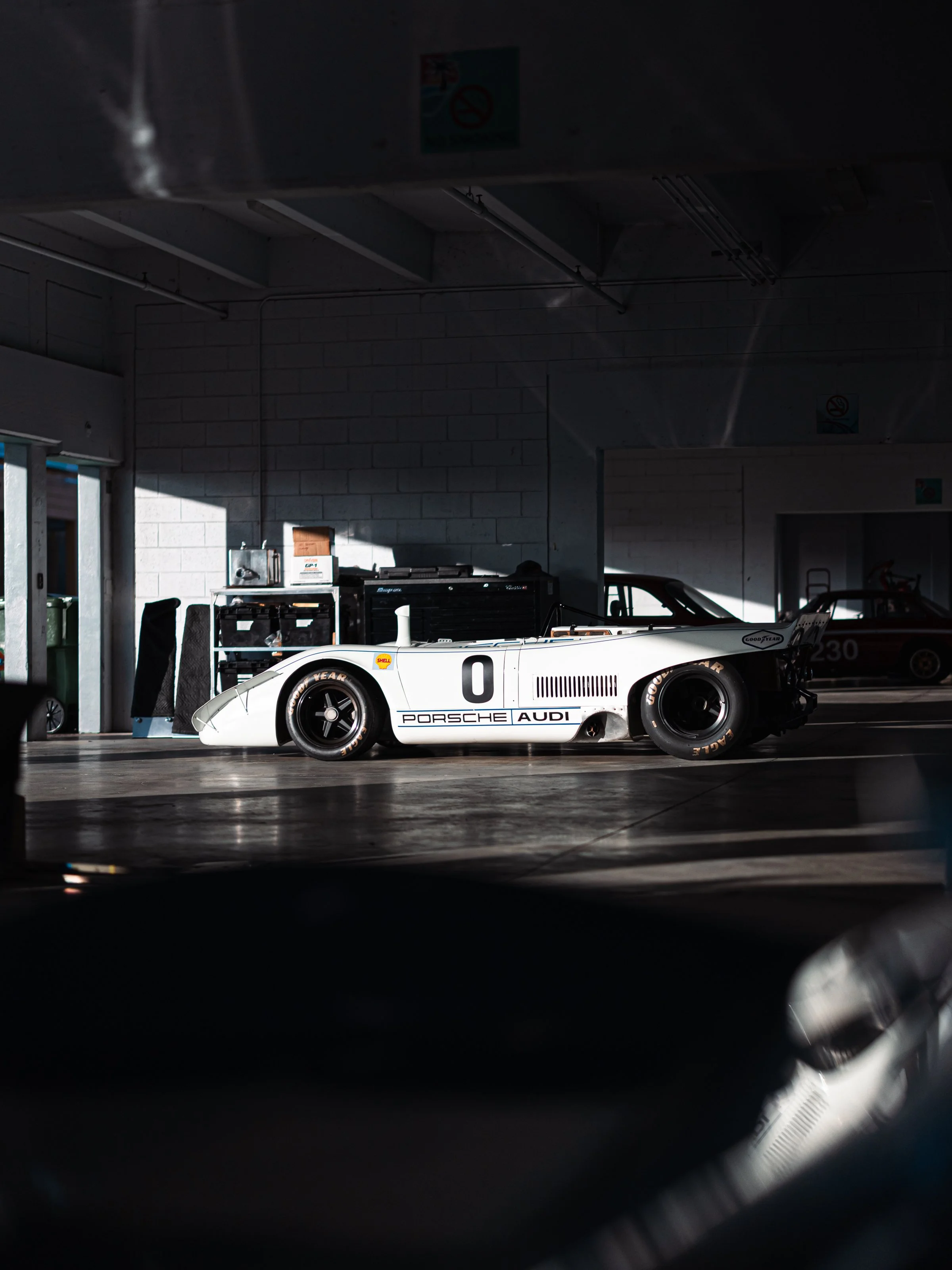 A vintage race car inside a garage with sunlight casting shadows on the wall.