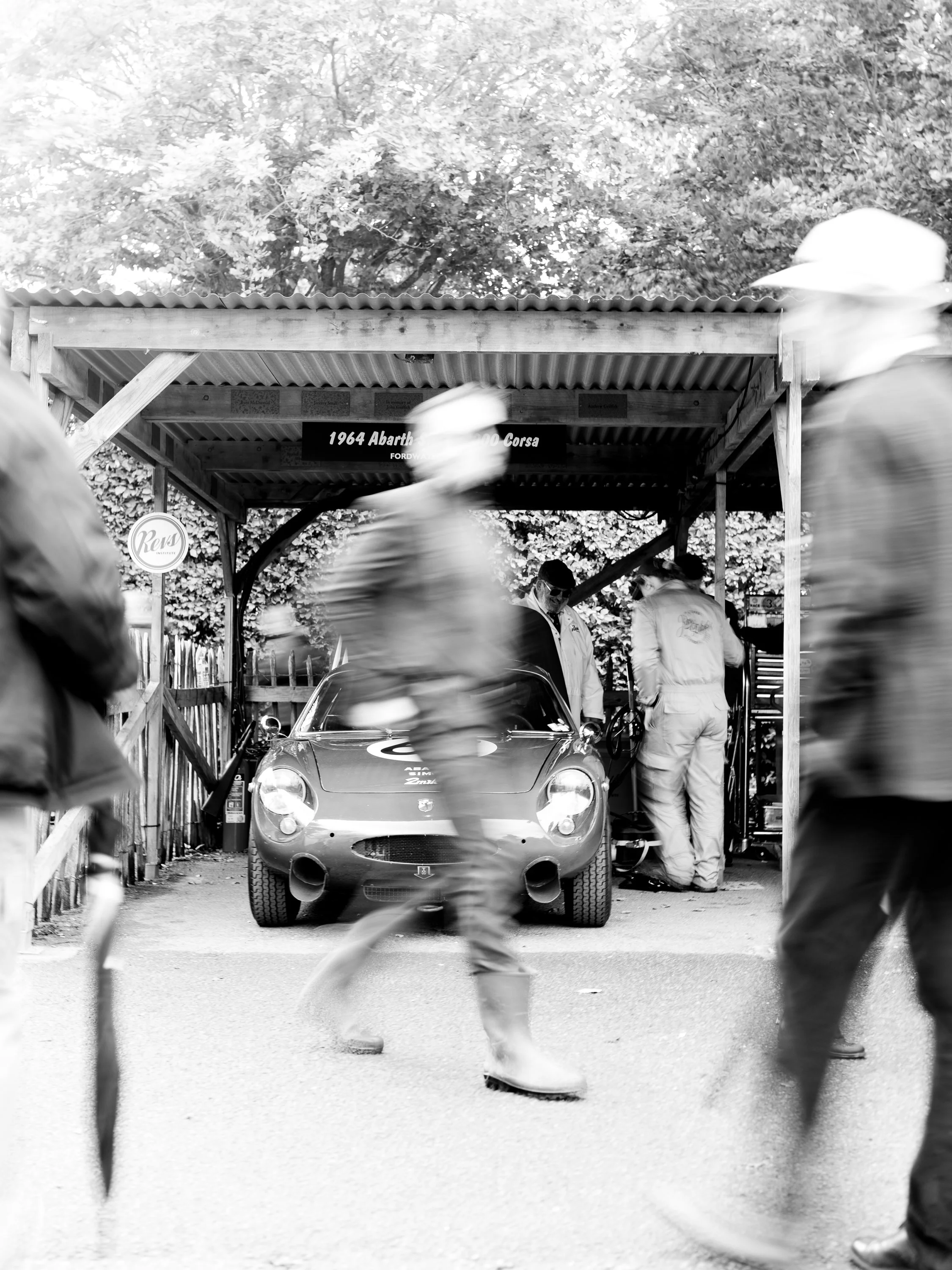Black and white photo of a classic racing car displayed in a wooden shelter with people walking by, some blurry due to motion.