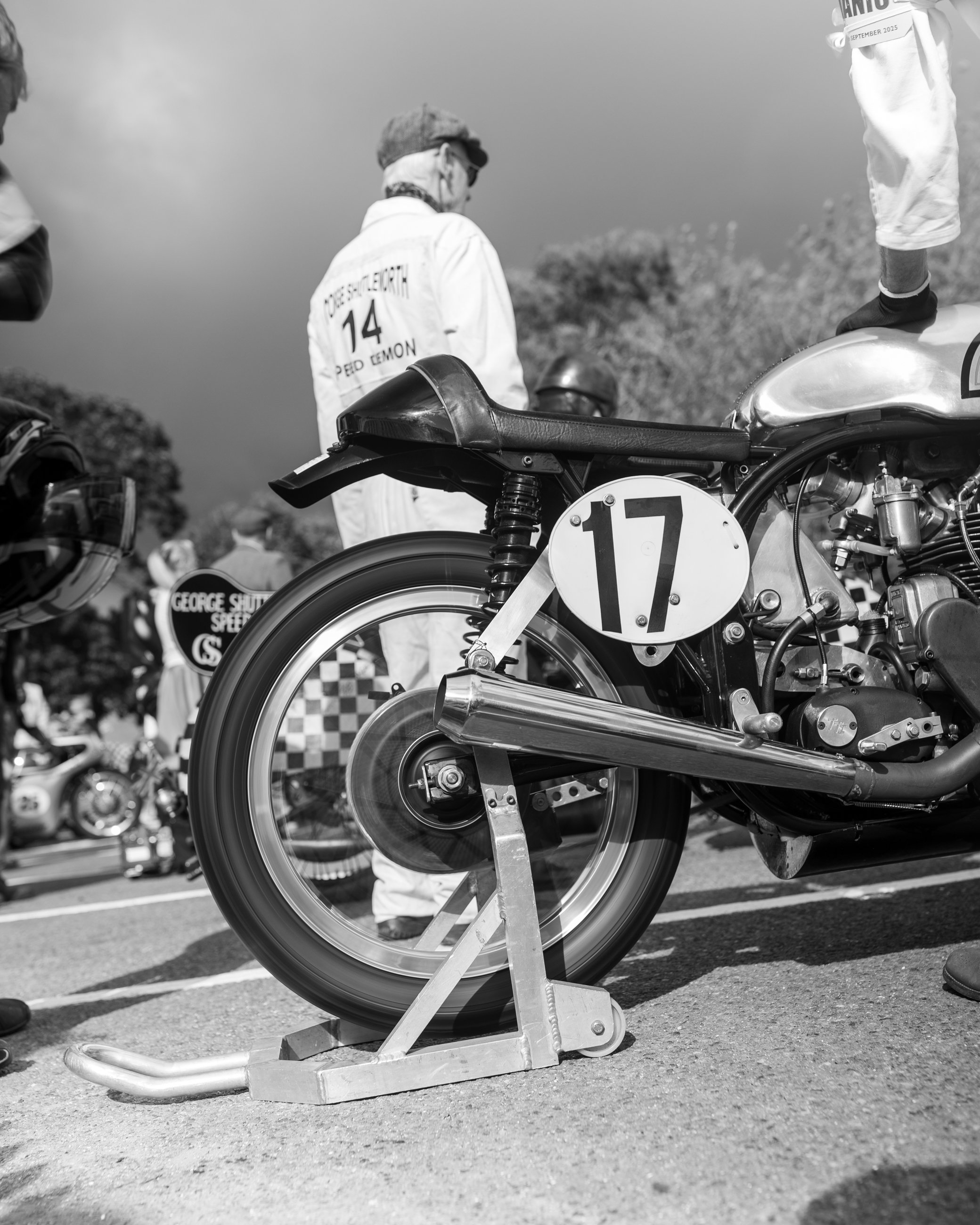 A vintage racing motorcycle on a stand at an outdoor race event with a rider in a helmet nearby. The motorcycle has a number 17 on its side and is surrounded by other bikes and people.