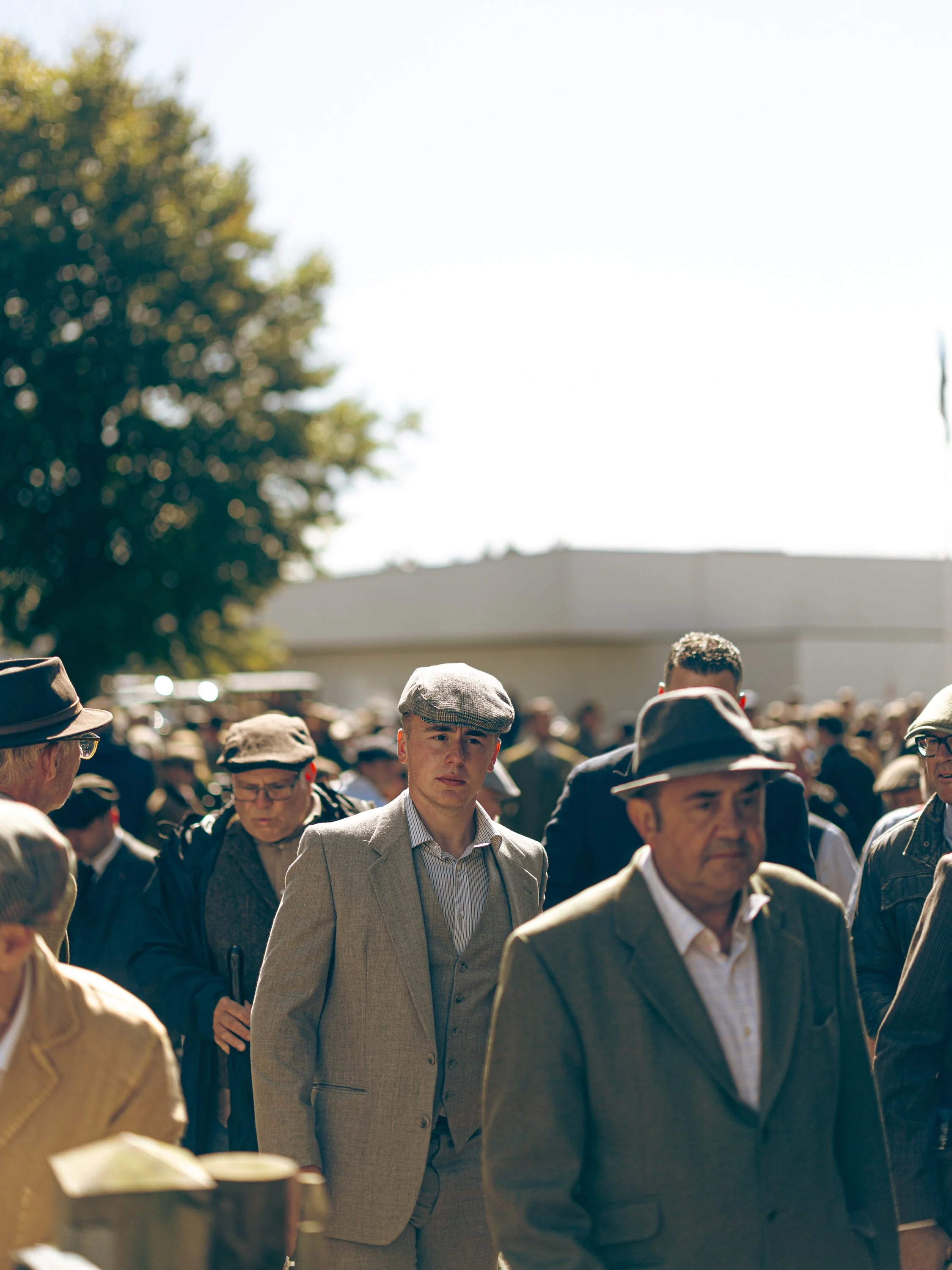Crowd of people walking outdoors on a sunny day, some wearing hats and suits, with a large tree and building in the background.