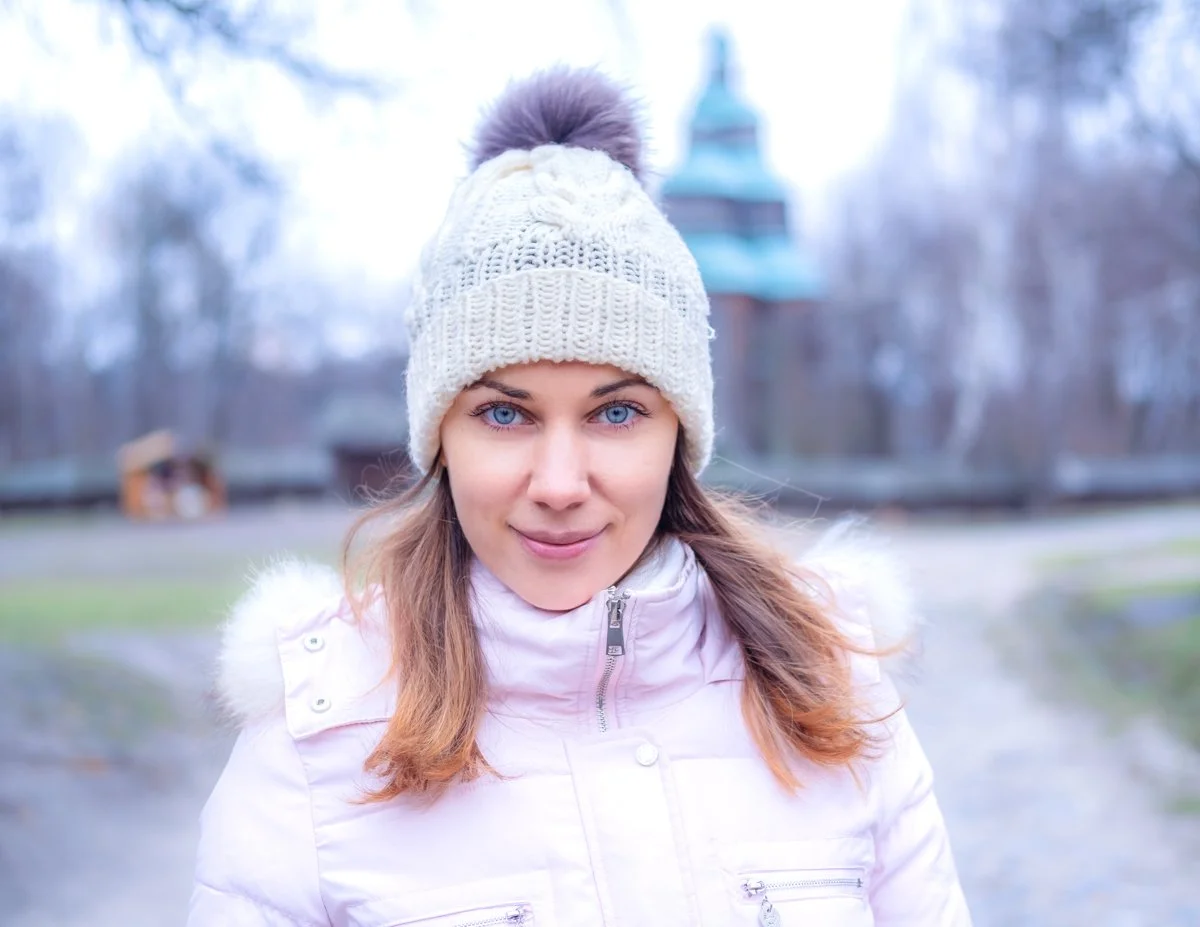 A young woman with blue eyes and light brown hair wearing a white knit hat with a pom-pom and a white winter jacket outdoors in a park during winter.