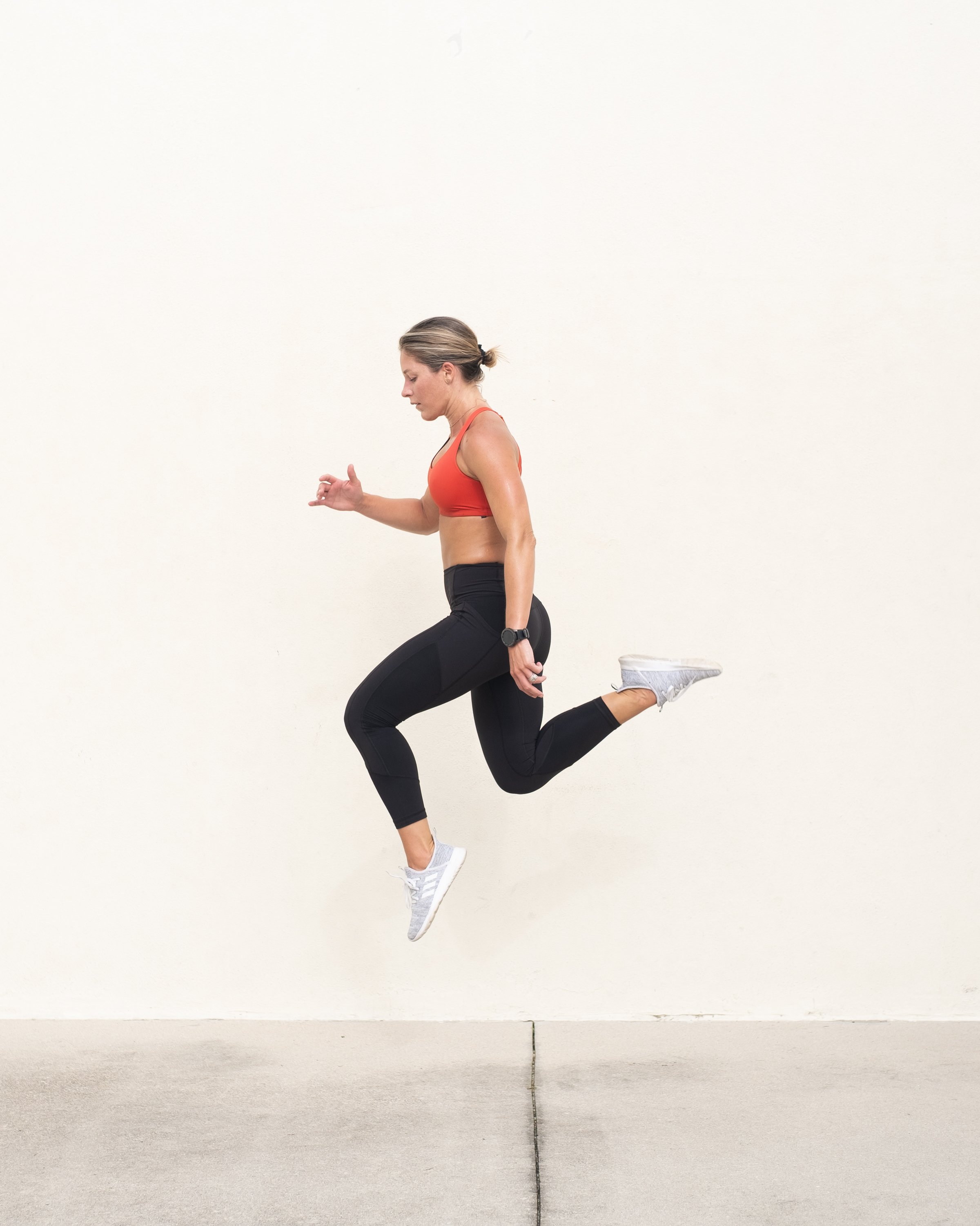 A woman in athletic clothing performing a lunge squat exercise while looking down, against a plain white wall.