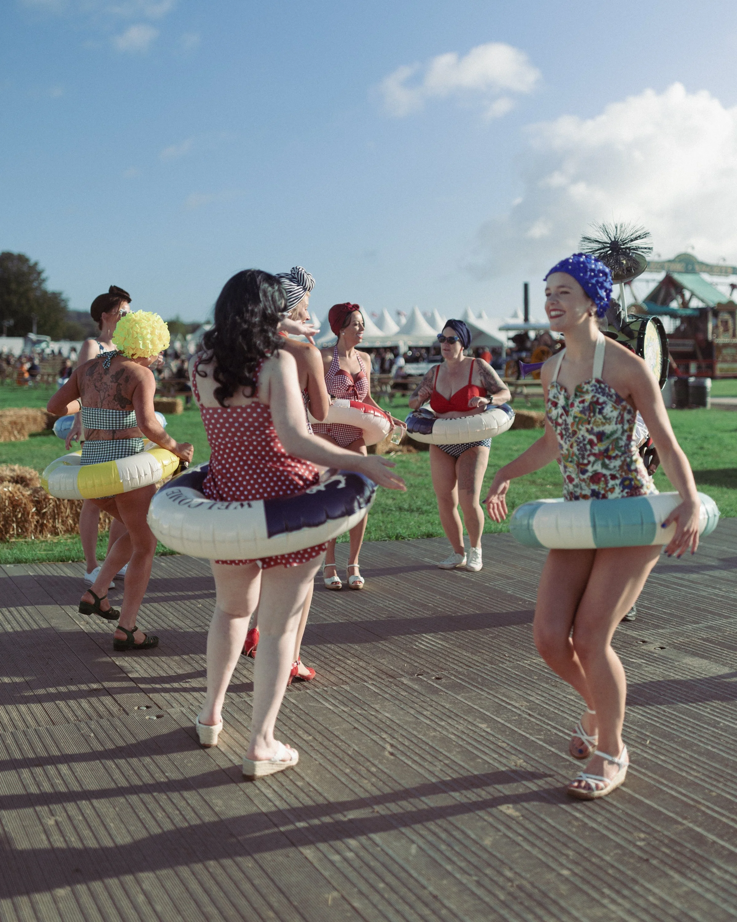 Women in vintage swimsuits and headscarves dancing and socializing outdoors on a sunny day.