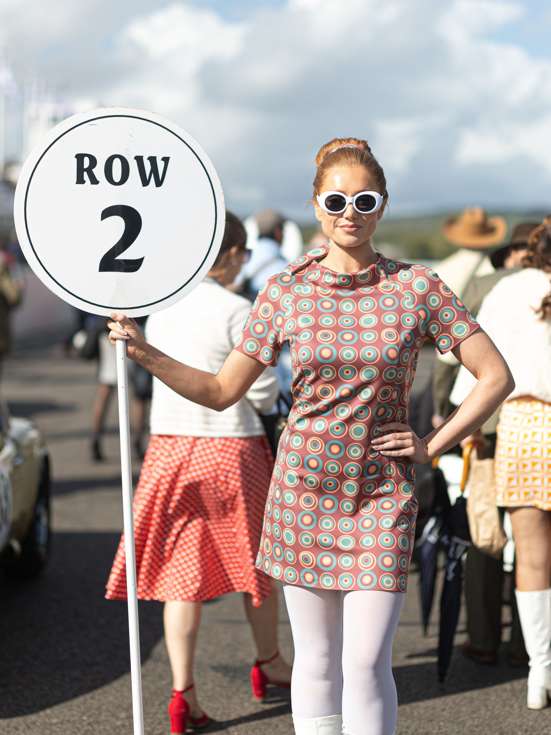 A woman wearing a vintage 1960s style dress with a geometric pattern, white tights, and large white sunglasses holding a sign that reads 'ROW 2'. She stands outdoors among other people, some dressed in retro or vintage clothing, under a partly cloudy