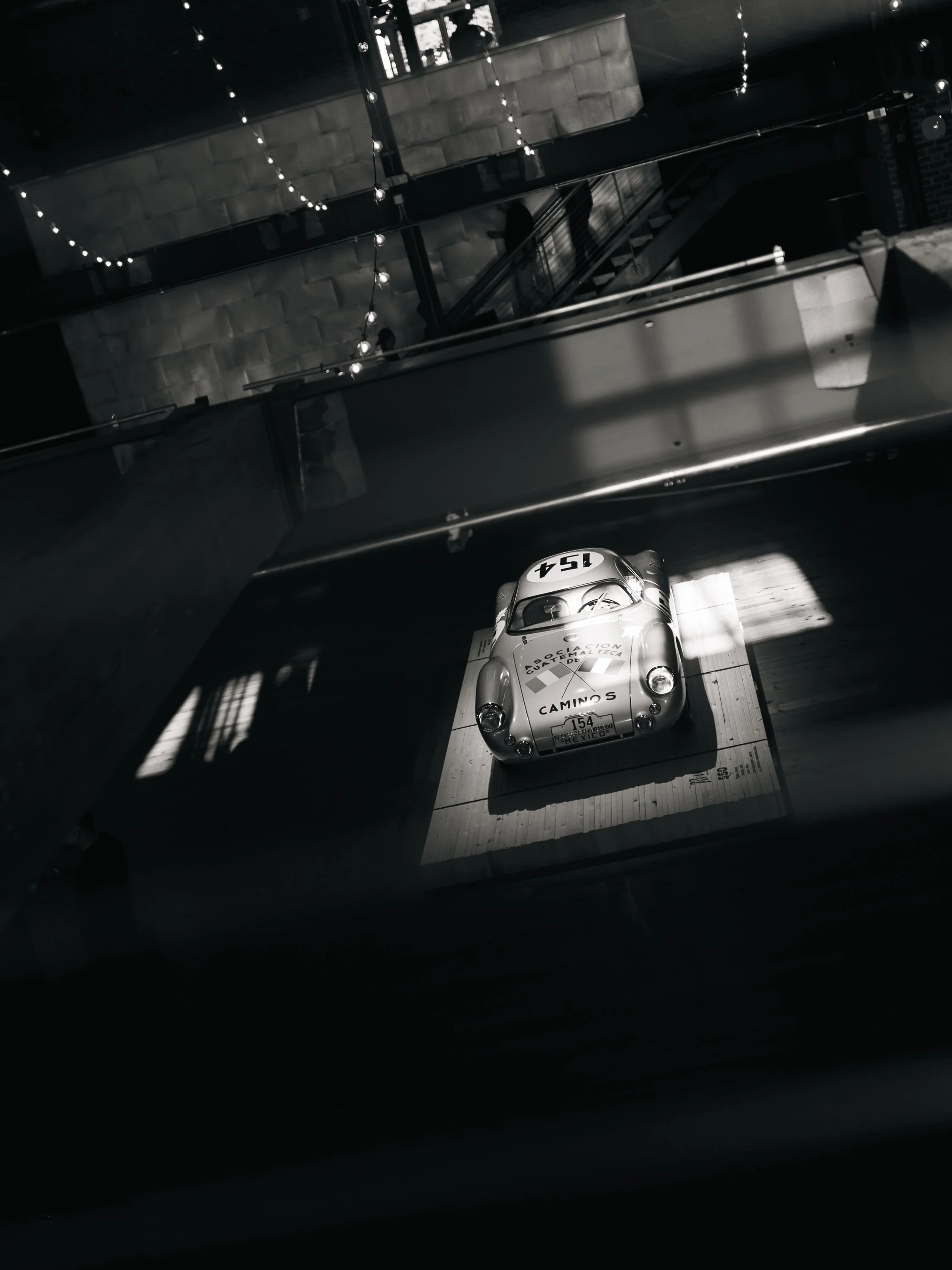A black-and-white photo of a vintage race car with the number 154 on its hood, displayed indoors on a platform near a glass railing with string lights hanging overhead.