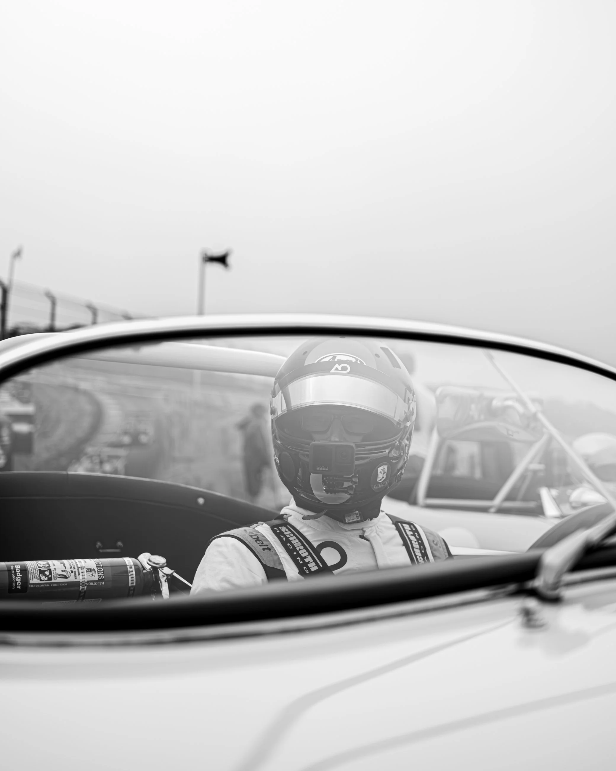 A race car driver wearing a helmet and racing suit sitting in a vehicle, with a race track and a flag in the background.