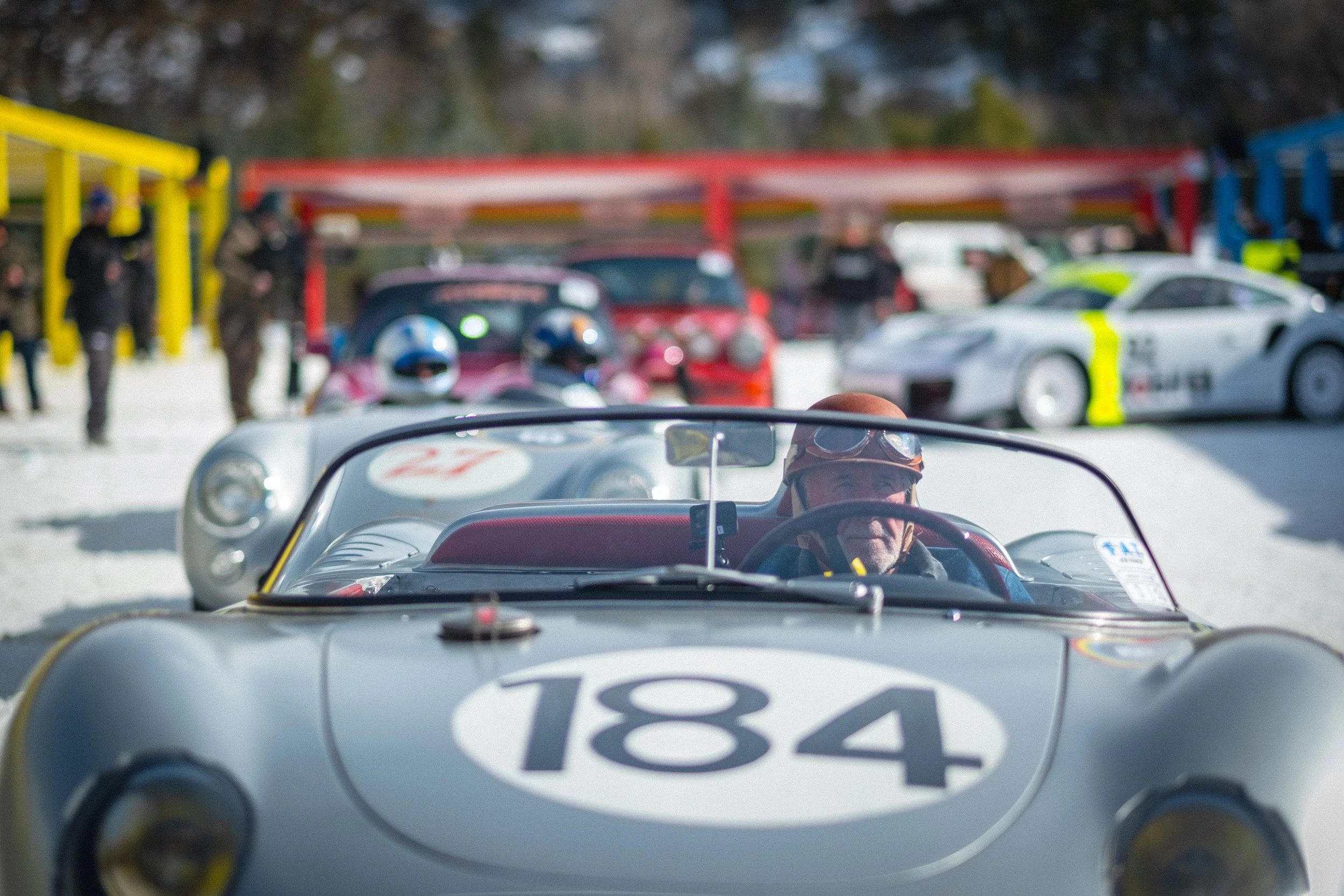 A vintage race car with the number 184 on the hood, driven by a person wearing goggles, with a line of other race cars, safety personnel, and spectators in the background at a snowy racetrack. Jeff Zwart, Porsche, Fat Ice Race, Aspen, Colorado