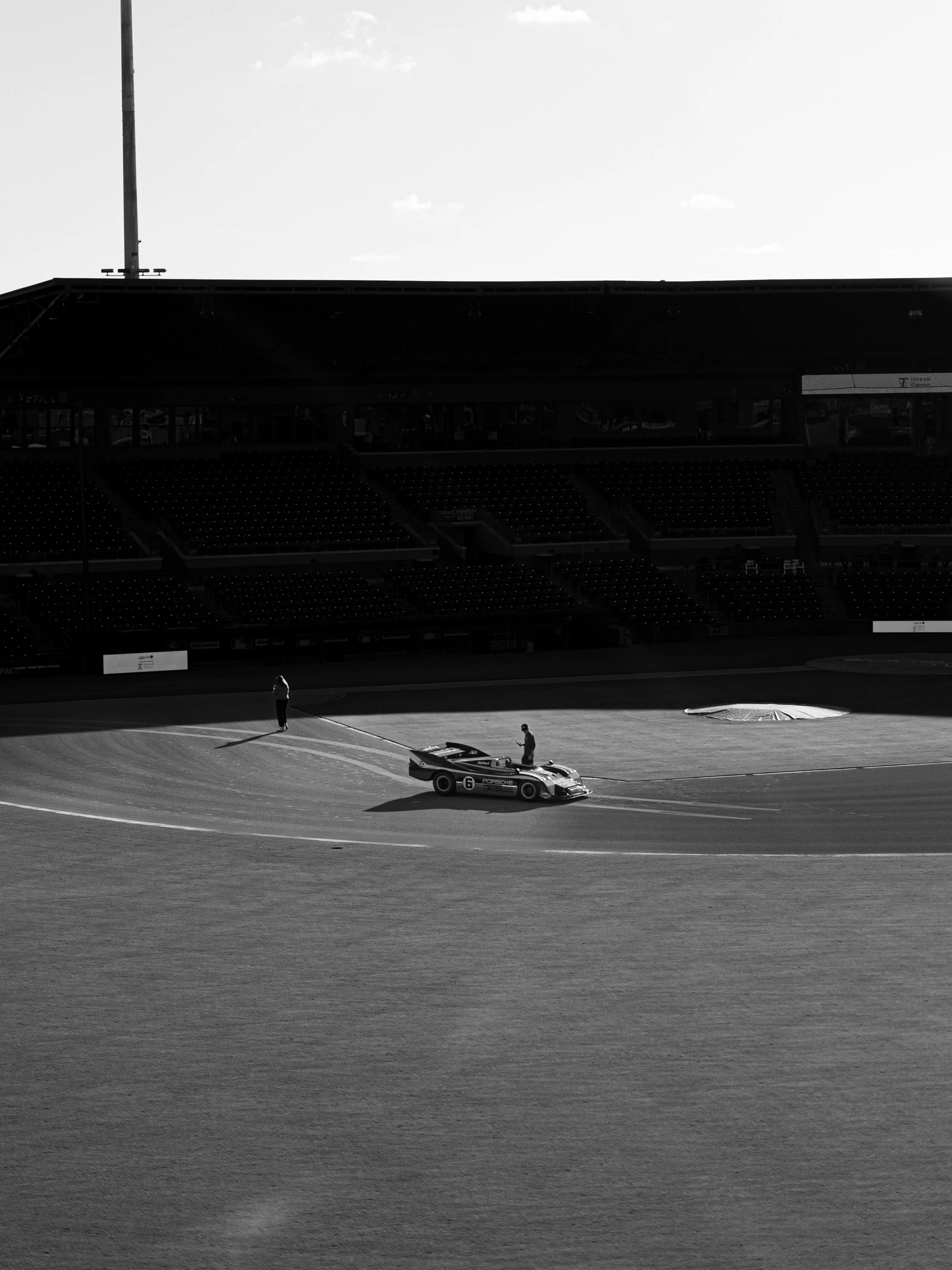 A black and white photograph of a racetrack with a Porsche racing car and two individuals next to it, under a stadium bleacher with a bright sky in the background.