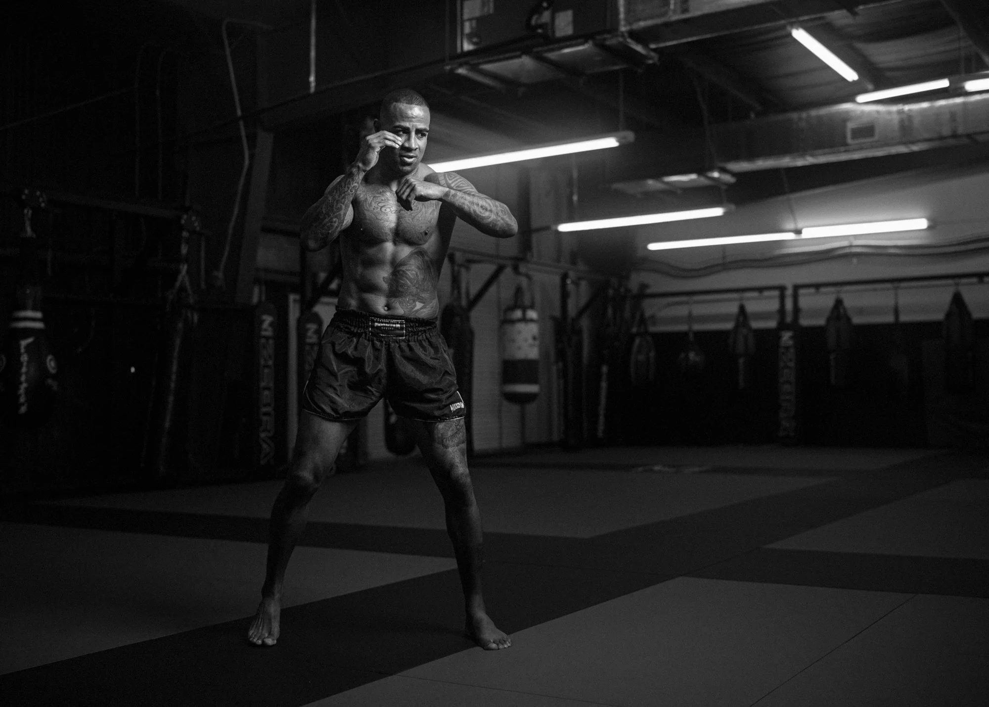 A shirtless male athlete with tattoos on his arms and chest stands in a gym, seemingly in a fighting stance, with one hand near his face and the other near his chest. The gym has boxing bags and exercise equipment in the background, and is lit with f