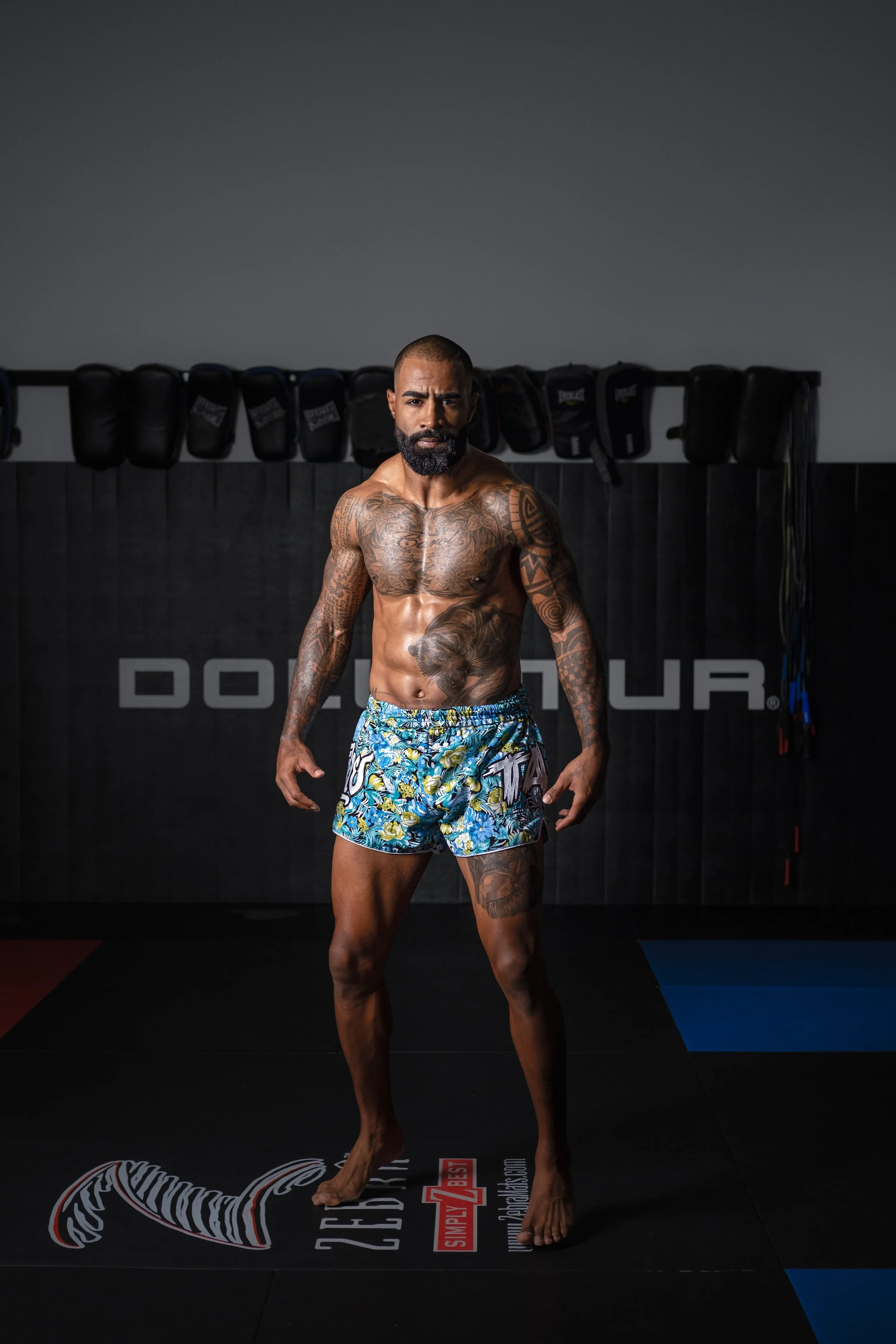 A muscular man with tattoos on his chest and arms, standing barefoot on a black and blue gym mat, wearing blue floral shorts, inside a gym with black boxing gloves hanging on the wall behind him. Cosmo Alexandre, muay thai