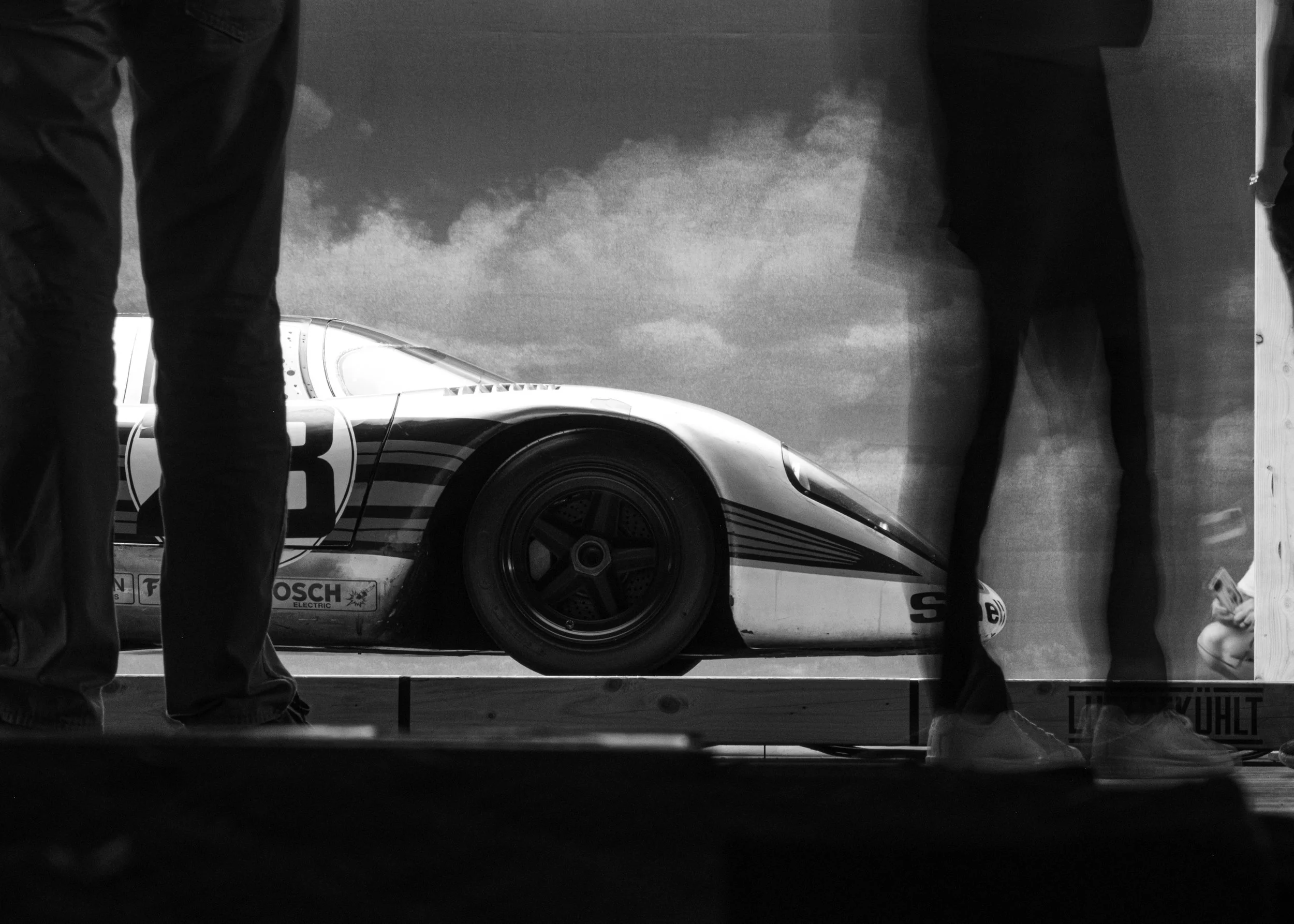 Black and white photo of two people standing near a race car, with only their legs visible, standing on a wooden platform. luftgekuhlt