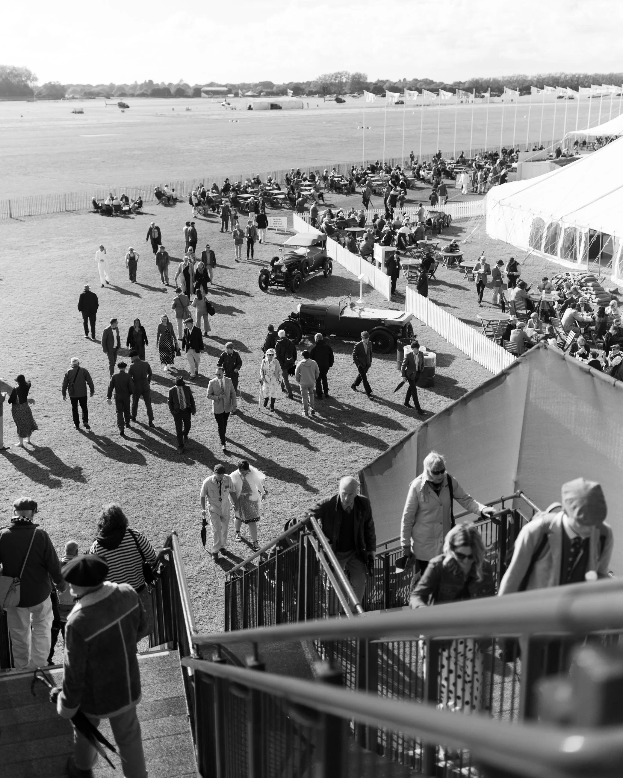 People walking up stairs and gathering outside a large tent, with a race track in the background.