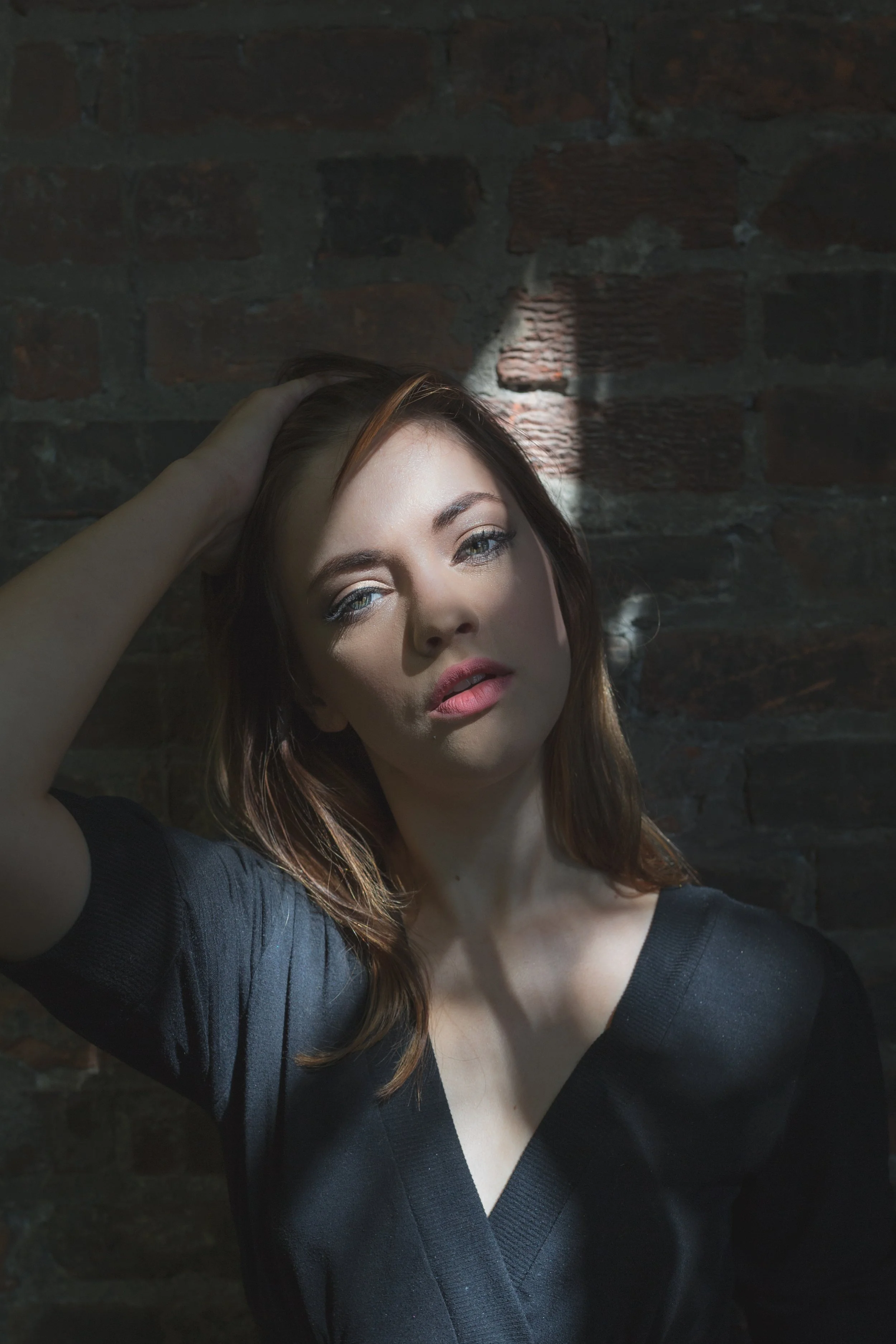 A woman with red hair and blue eyes standing in front of a brick wall, holding her head with one hand, wearing a black V-neck top, with sunlight casting shadows on her face.